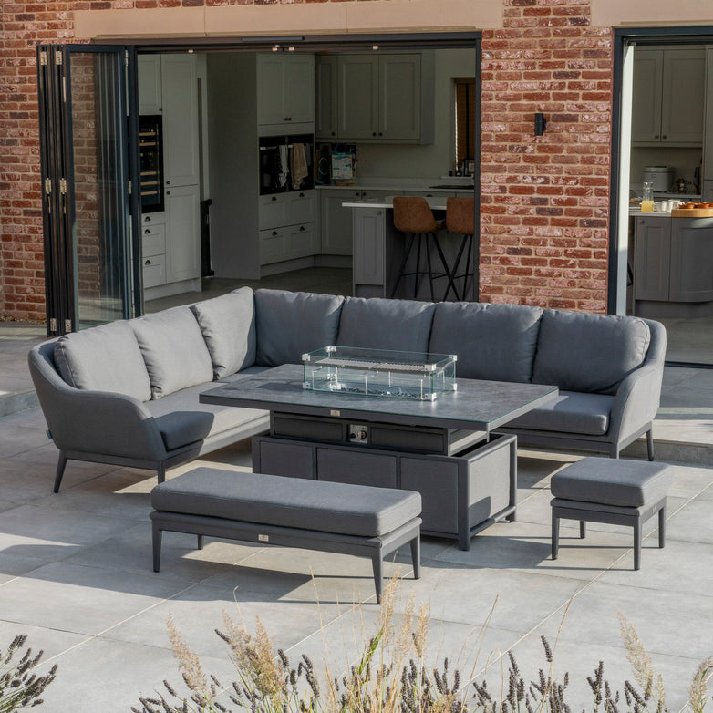 Outdoor patio featuring the Luna Outdoor Fabric Rectangular Corner Dining Set with Rising Firepit Table in Grey (Left Hand), set in front of a modern brick home with open doors.