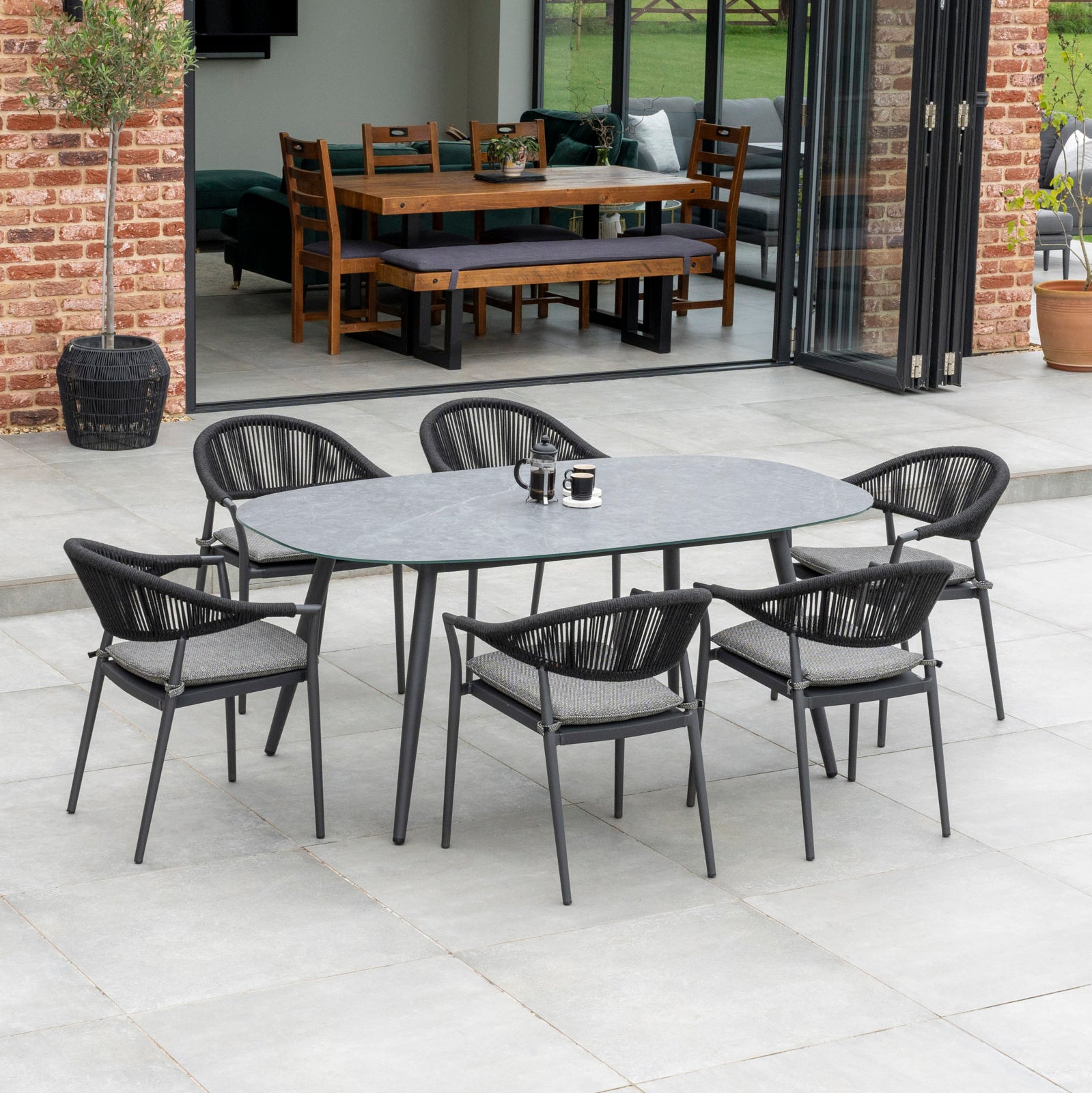 Outdoor patio with a Cloverly 6 Seat Rope Oval Dining Set with Ceramic Table in Charcoal, featuring six black woven chairs and a ceramic glass table. Two mugs sit on the table, while the indoor dining area is visible through open sliding doors.