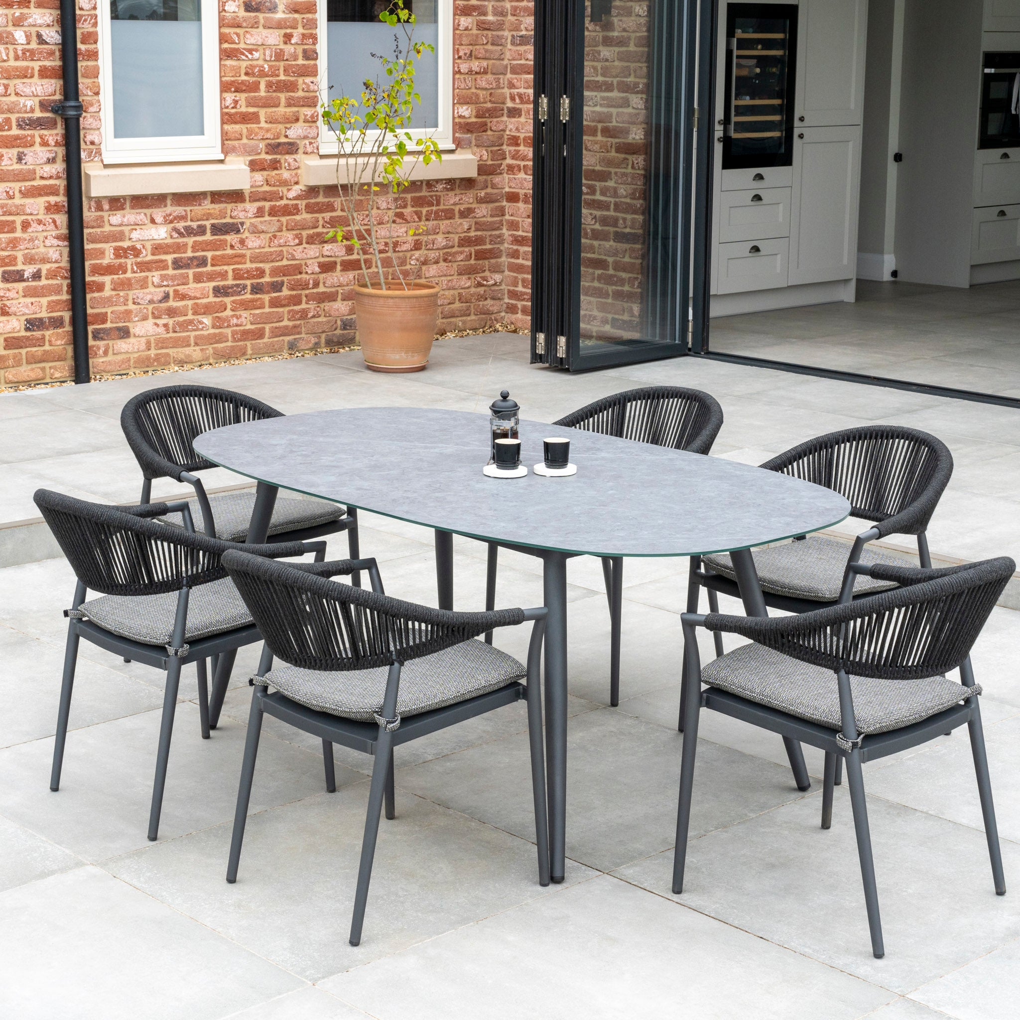 Outdoor dining area with the Cloverly 6 Seat Rope Oval Dining Set featuring a ceramic table in charcoal, six woven chairs, and two coffee cups. Brick wall and open sliding doors in the background.
