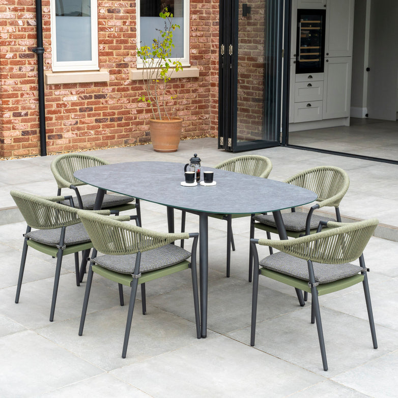 Outdoor dining area featuring the Cloverly 6 Seat Rope Oval Dining Set with a Ceramic Table in Olive Green, complemented by woven chairs with green cushions, set on a stone patio with a brick wall backdrop.