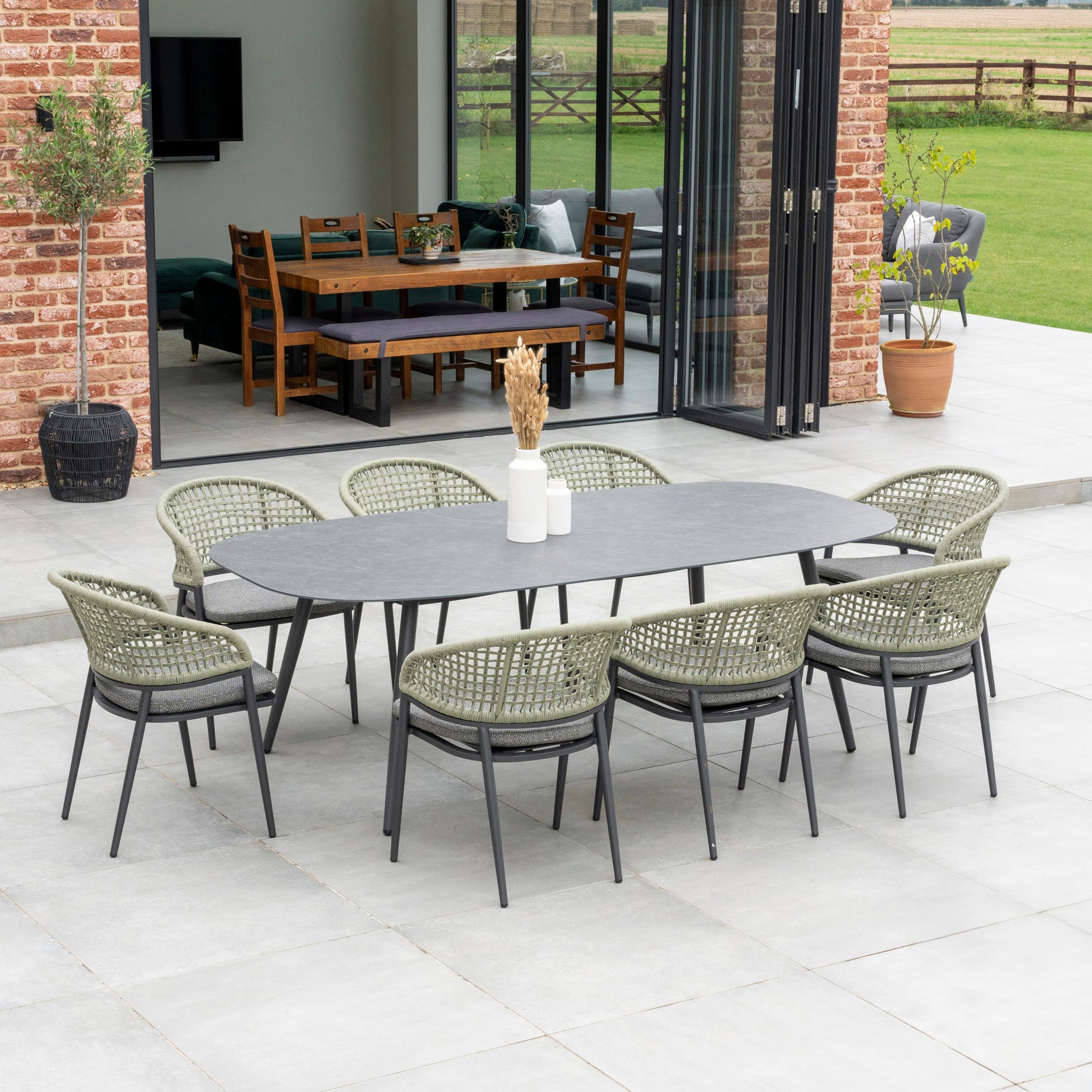 The outdoor patio features the Kalama 8 Seat Rope Oval Dining Set in Olive Green, with eight exquisitely woven chairs surrounding a weatherproof ceramic table, offering a seamless view into the inviting living area.