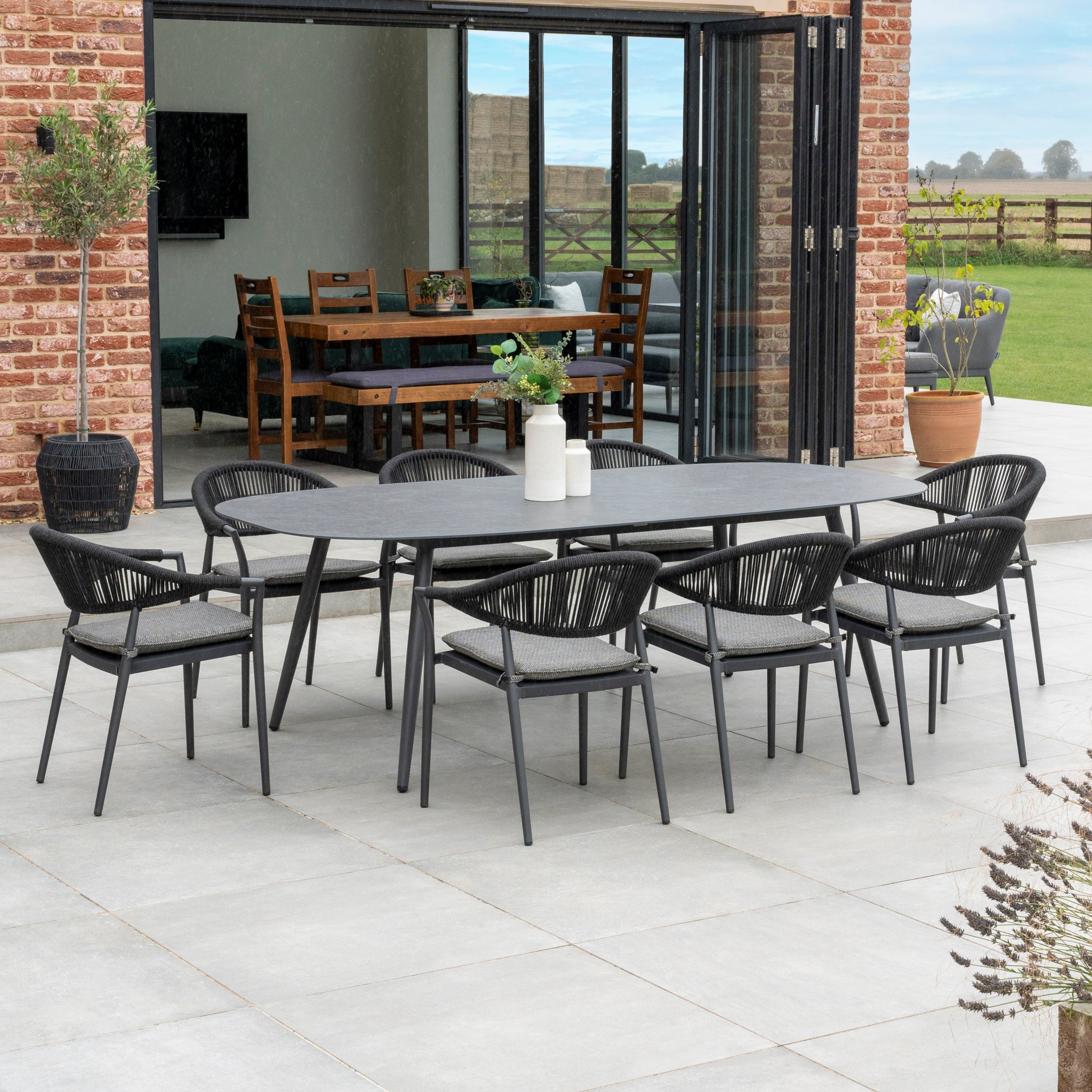 The Cloverly 8 Seat Rope Oval Dining Set with Ceramic Table in Charcoal features a maintenance-free aluminum frame and includes a long, ceramic glass table surrounded by eight black chairs. This outdoor dining area is located on a patio with a beautiful view into the modern house.