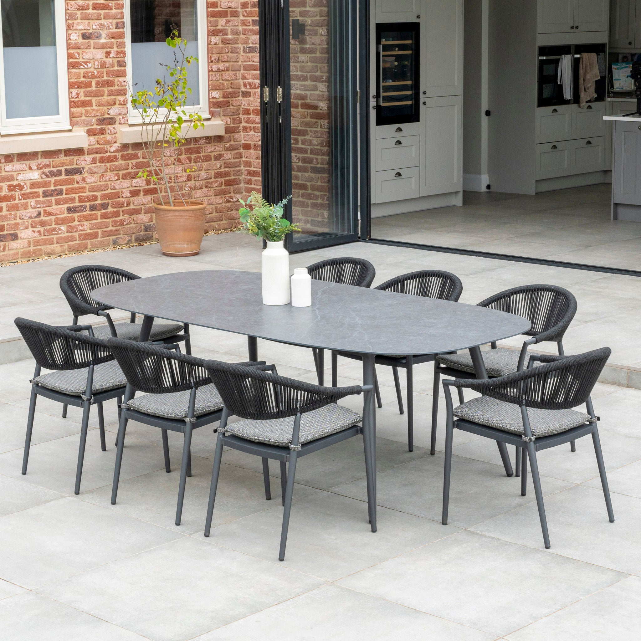 The Cloverly 8 Seat Rope Oval Dining Set with a Ceramic Table in Charcoal, featuring a maintenance-free aluminum frame, is elegantly arranged on the patio of a modern house with an open sliding door.
