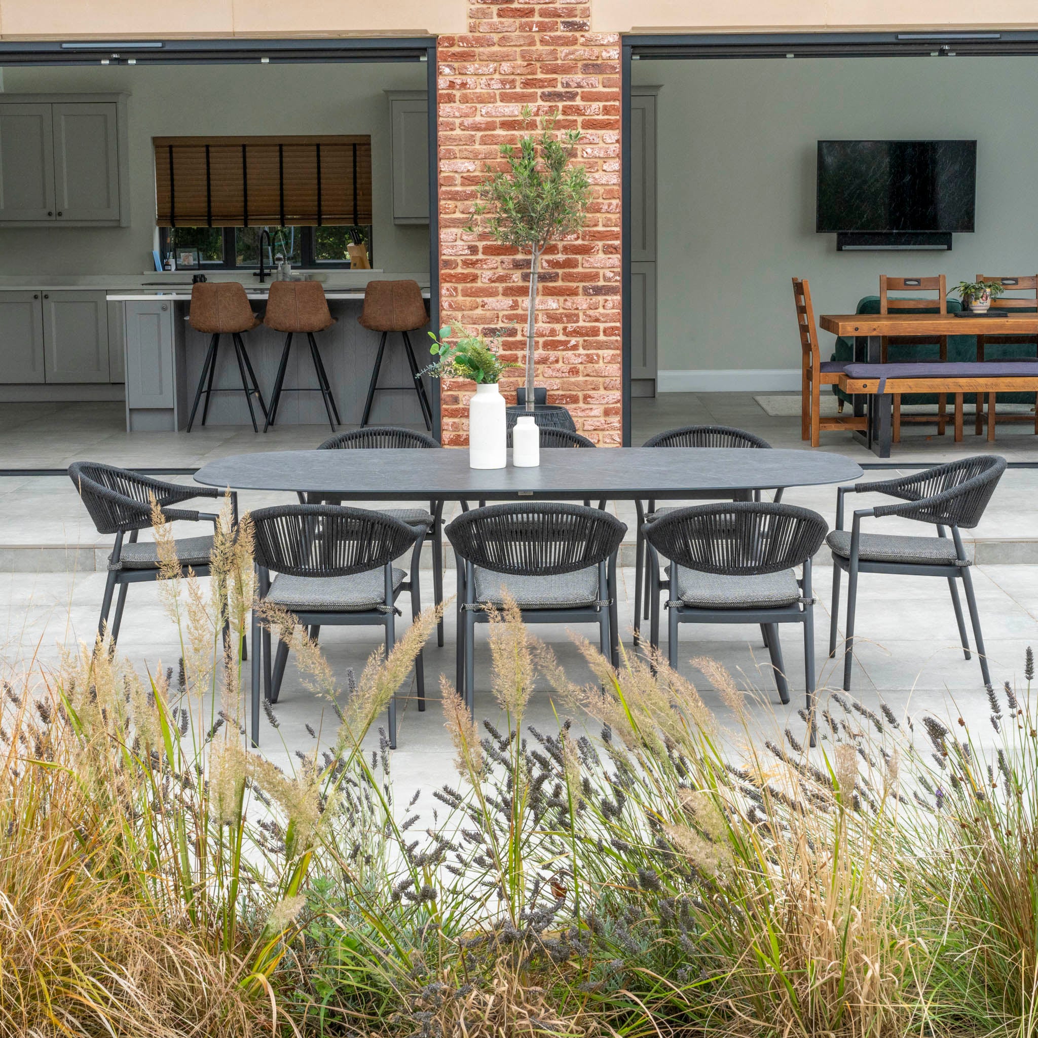 Outdoor dining area featuring the Cloverly 8 Seat Rope Oval Dining Set with a sleek ceramic table in charcoal, complemented by eight chairs and decorative items, situated between open indoor kitchen and dining areas. Crafted from maintenance-free aluminum for ease and durability.