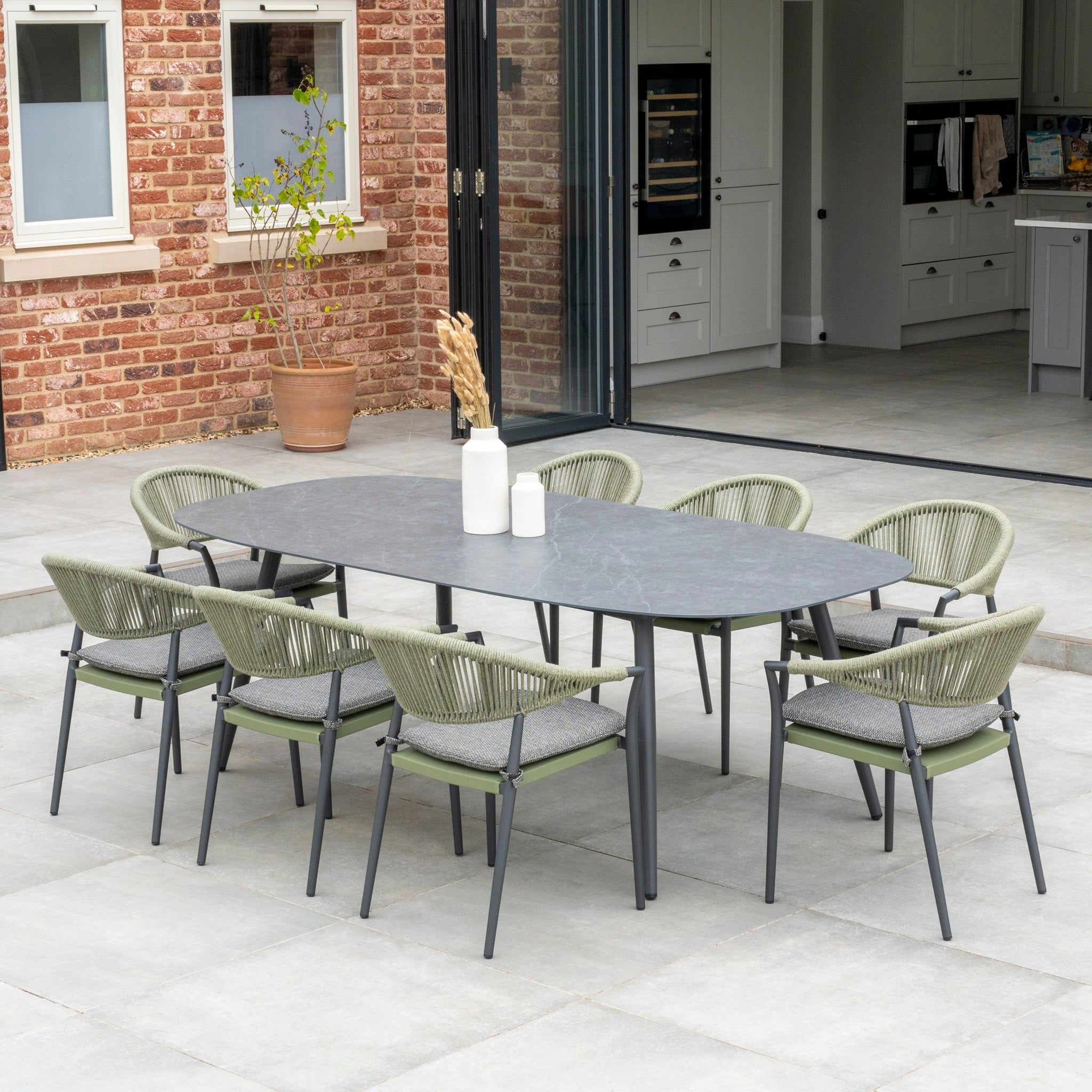 Outdoor patio with a Cloverly 8 Seat Rope Oval Dining Set in Olive Green, featuring a ceramic table and stackable woven chairs, set against a modern brick and glass house exterior.