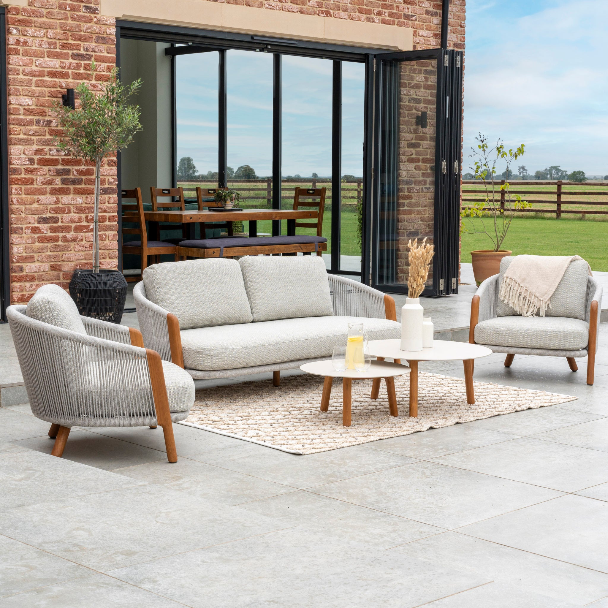 The outdoor seating area features the Merano 2 Seat Sofa Set and Coffee Tables in Latte, beautifully crafted with luxurious wicker and hydrophobic olefin rope weave. A rug and a round table adorn the patio, while glass doors reveal a view of the dining area inside.