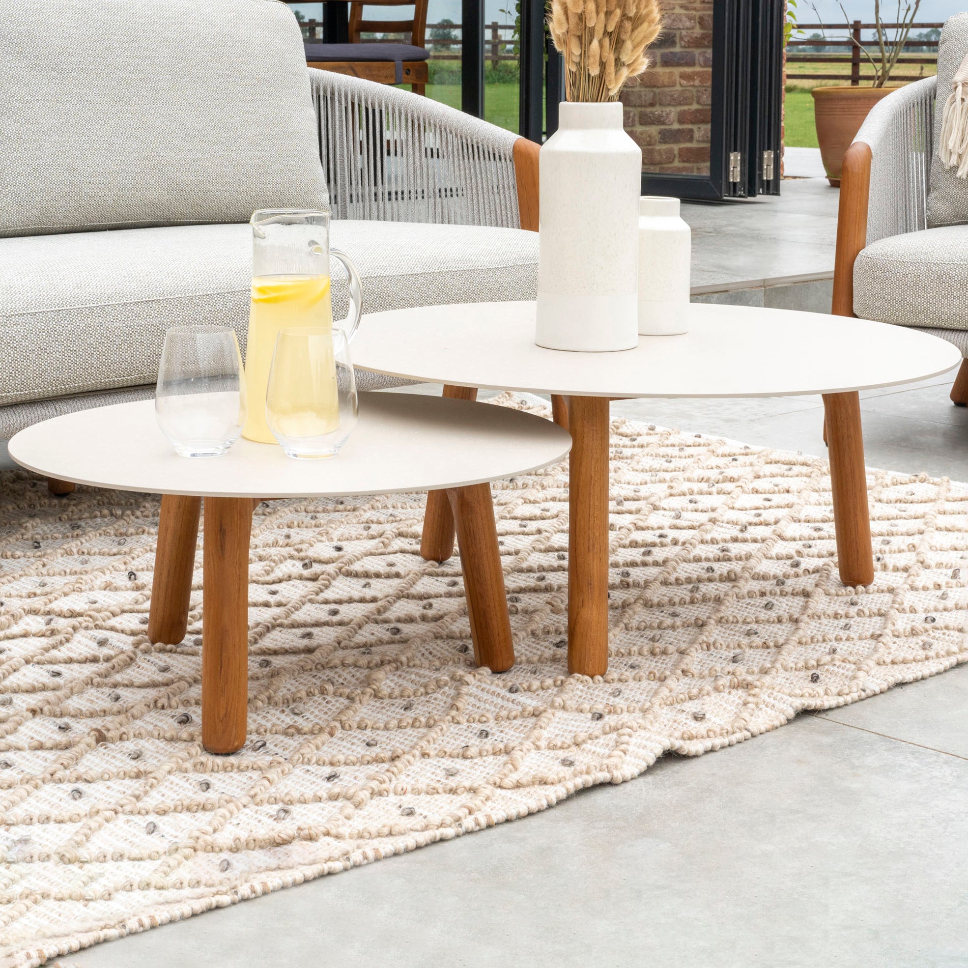 Two round wooden tables, one smaller, holding a glass pitcher of lemonade, two glasses, and decorative vases on a rug beautifully complement the Merano 2 Seat Sofa Set and Coffee Tables in Latte. This outdoor luxury furniture features hydrophobic olefin rope weave for added style and durability.