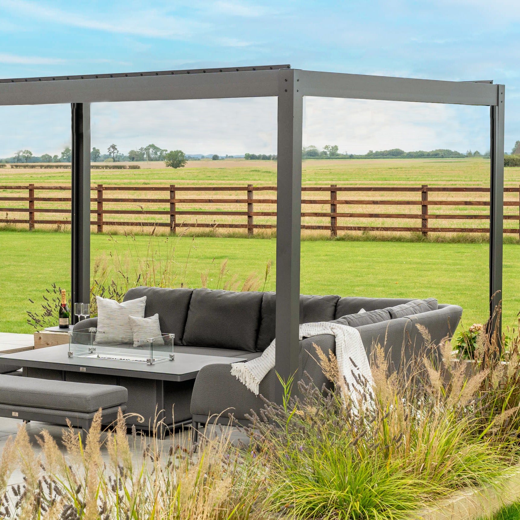 Modern outdoor seating area with gray furniture under a PergoSTET Plus 3m x 3m Square Pergola with LED Lighting – Frame Only in Grey, overlooking a grassy field and wooden fence.