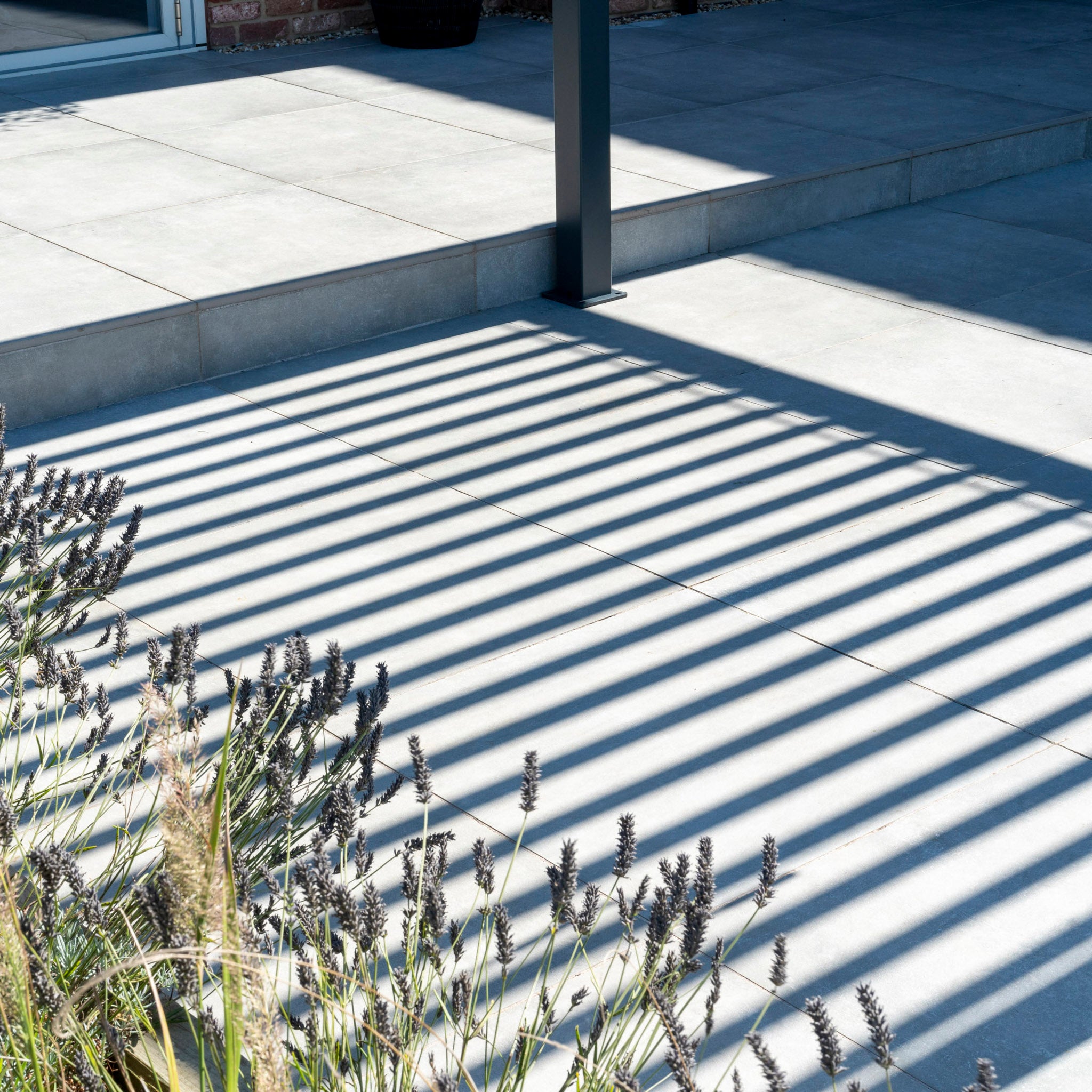 Shadows of the PergoSTET Plus 3m x 4m Rectangular Pergola with LED lighting and three drop sides waltz across a sunlit tiled patio, as lavender plants sway gracefully in the foreground.