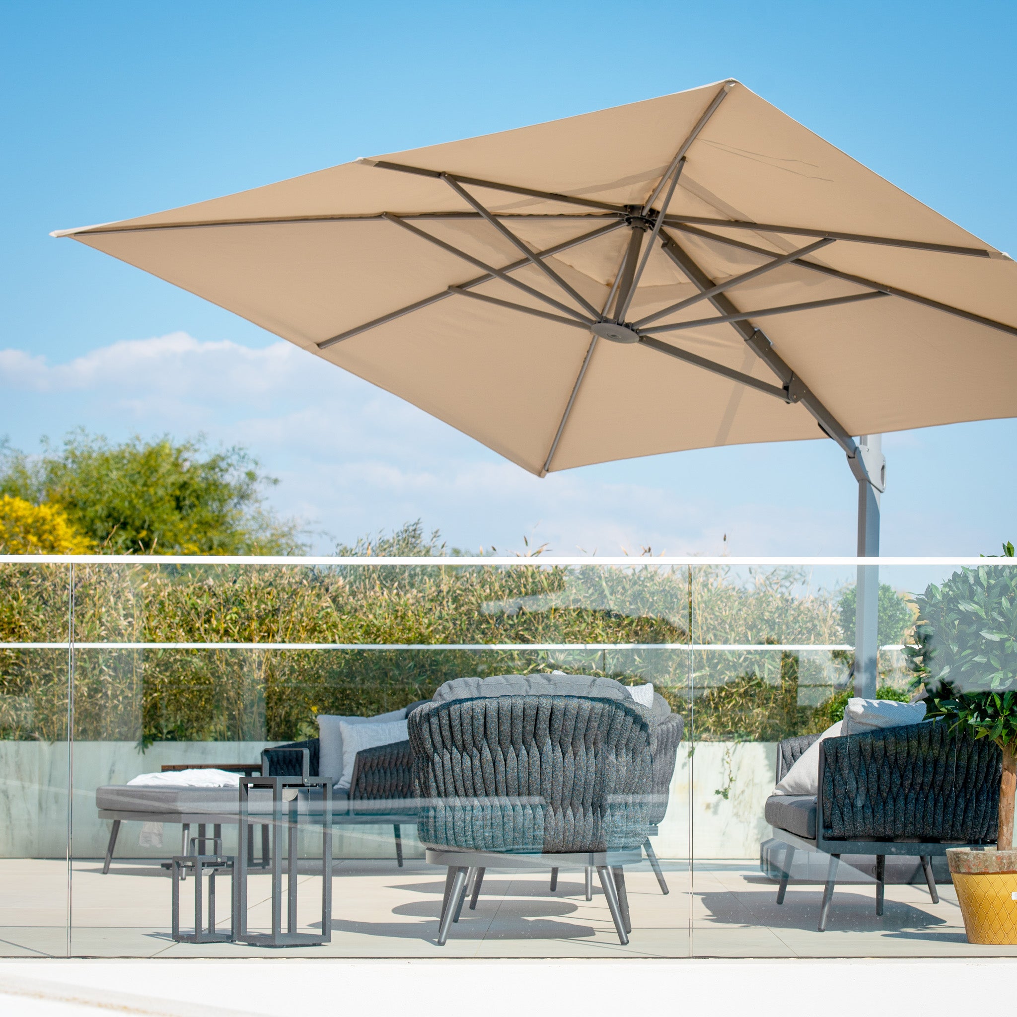 Outdoor patio seating with the Pallas 4m x 3m Rectangular Cantilever Parasol with LED Lighting in Beige, glass railing, and lush greenery under a clear sky—perfect for relaxing in elegant garden shade.