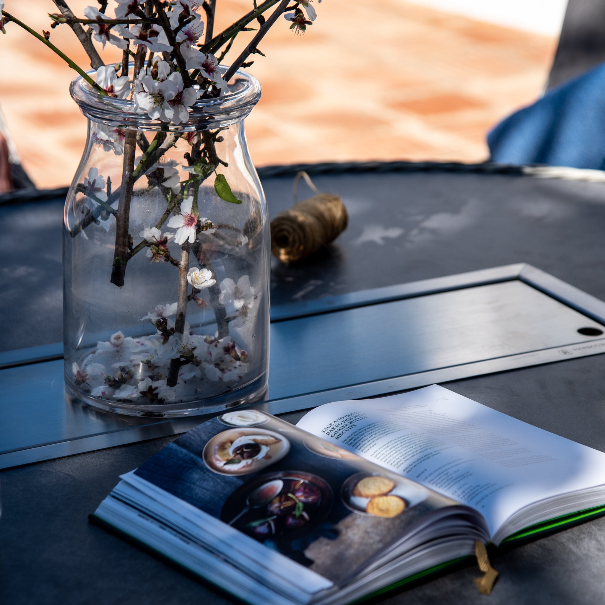 A glass jar with flowering branches beside an open book rests on a Santiago 180cm x 120cm Firepit Oval Dining Table in White Wash, illuminated by sunlight.