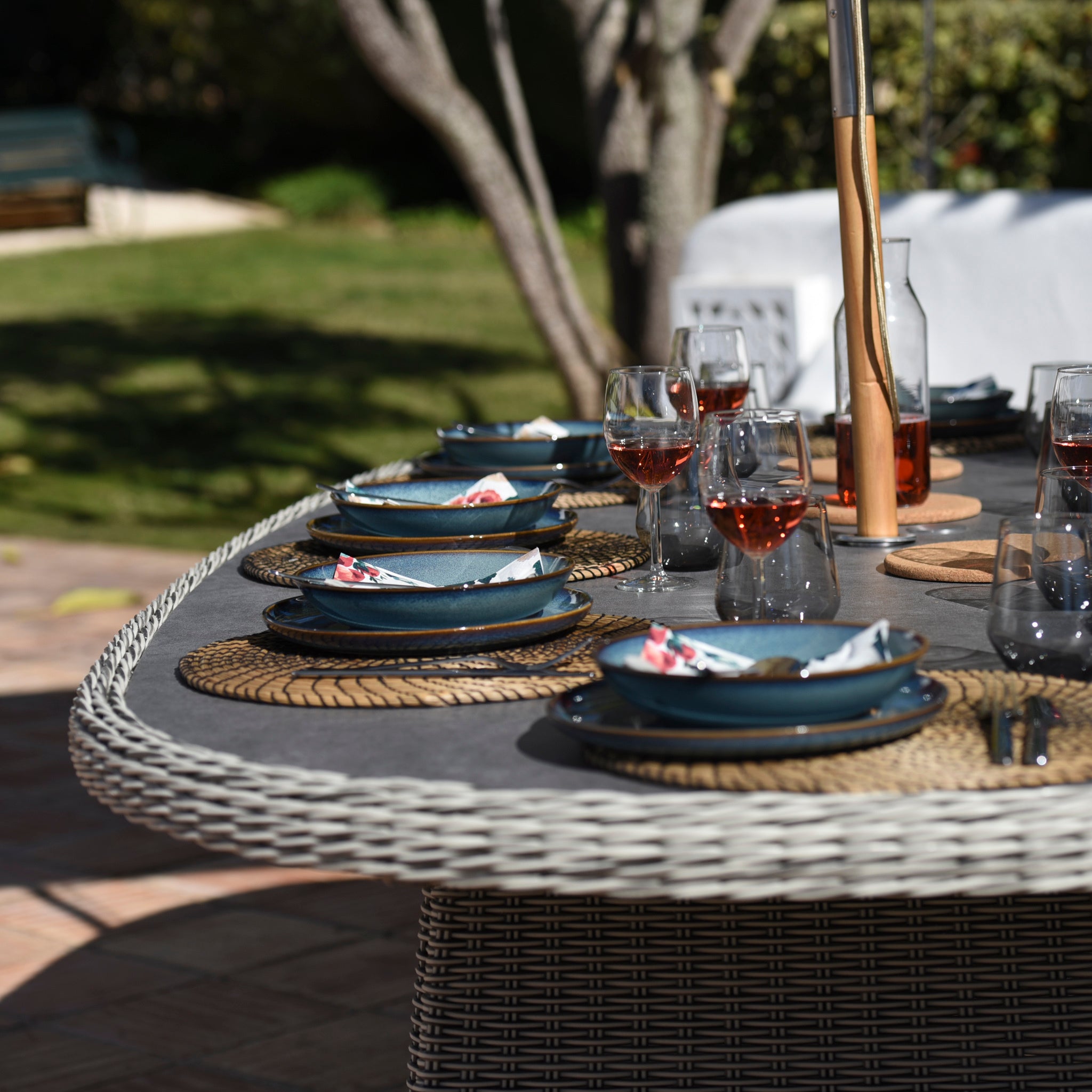 The Santiago 180cm x 120cm Firepit Oval Dining Table in White Wash is ideal for sunny days, featuring plates, napkins, and glasses of rosé—perfect outdoor furniture for relaxing meals.