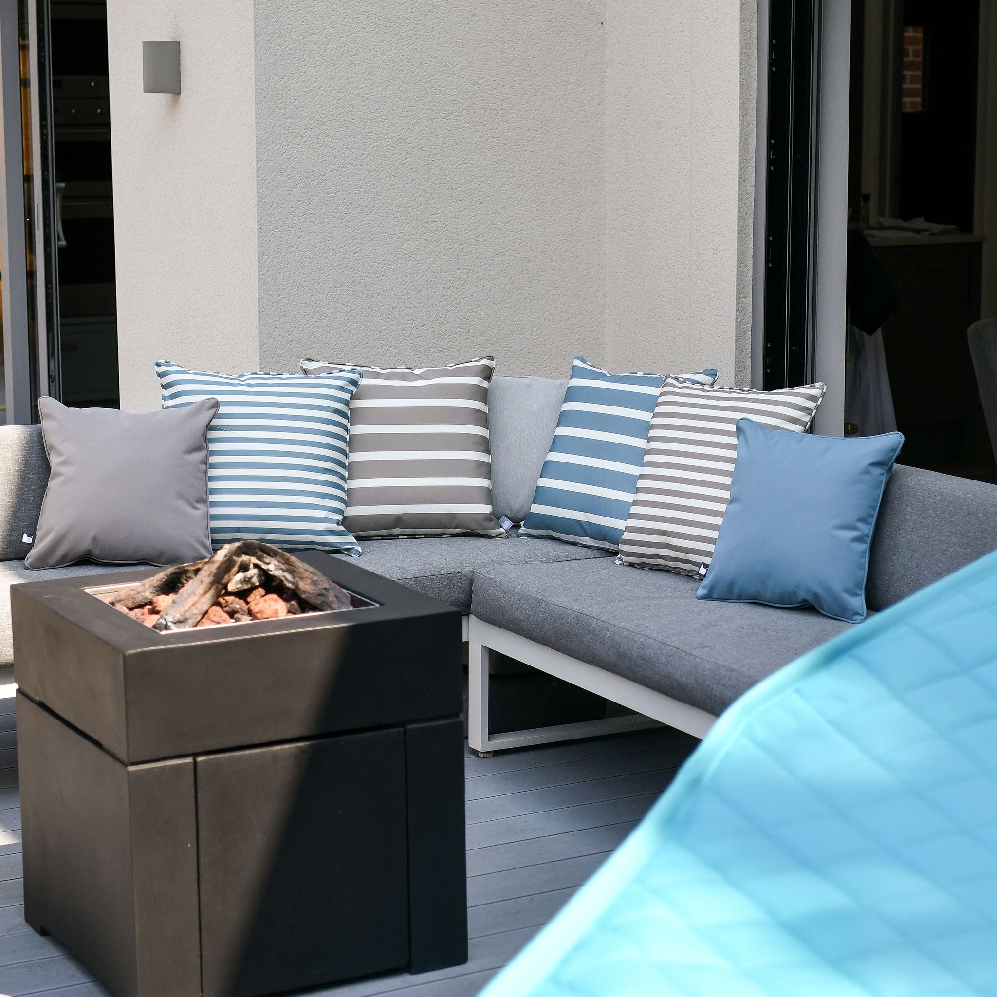 Outdoor seating area with a gray corner sofa, Outdoor Awning Stripe B-Cushion in Sea Blue, plain blue B-bag cushions, breathable polyester fabric for ultimate comfort, and a square fire pit table.