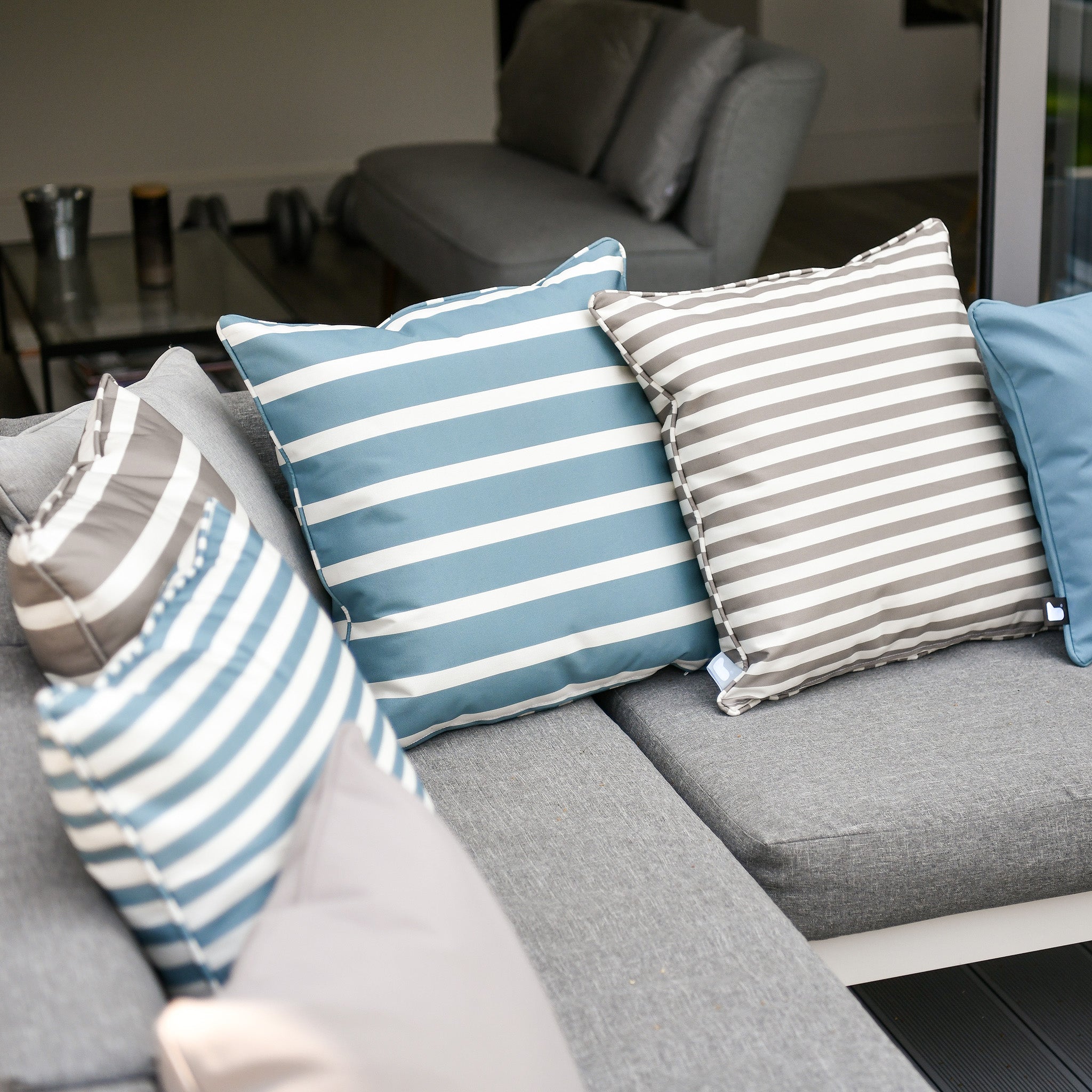 Outdoor seating area featuring UV-resistant, breathable Outdoor Awning Stripe B-Cushion in Sea Blue, accented by blue, gray, and white striped pillows, alongside a table with decor.