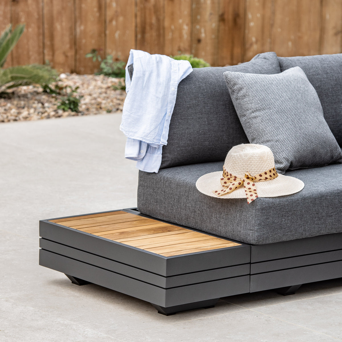 The Panama Teak Side Table sits alongside a gray outdoor sofa with an aluminium frame, complemented by a straw hat and polka dot scarf, plus a white shirt draped over the back—an inviting scene of stylish outdoor furniture.