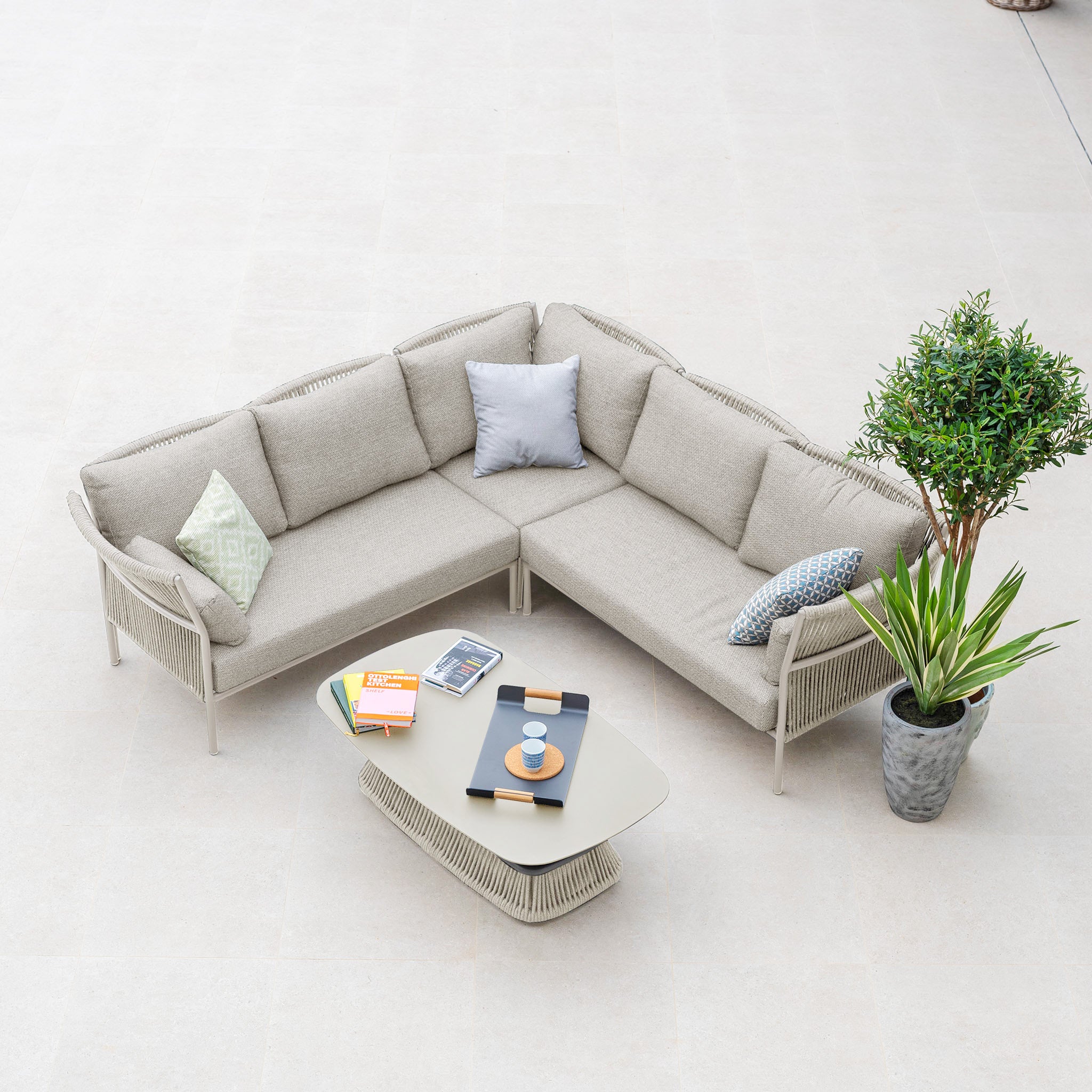 The Salina Corner Group Set with Aluminium Table in Latte, a weather-resistant outdoor furniture set, features an L-shaped beige sofa with cushions, a sleek aluminium table adorned with books and cups, and two potted plants on a light floor.