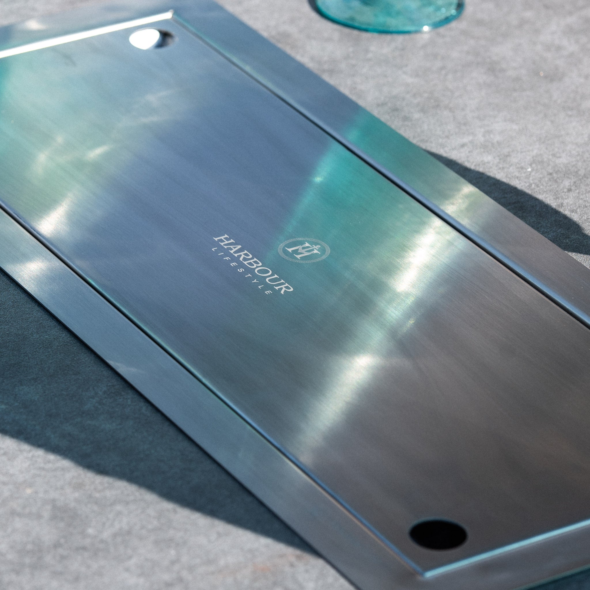 Rectangular stainless steel tray with "Harbour Lifestyle" logo on a gray surface beside a glass—an elegant accessory that perfectly complements the Santiago 180cm x 120cm Firepit Oval Dining Table in White Wash.