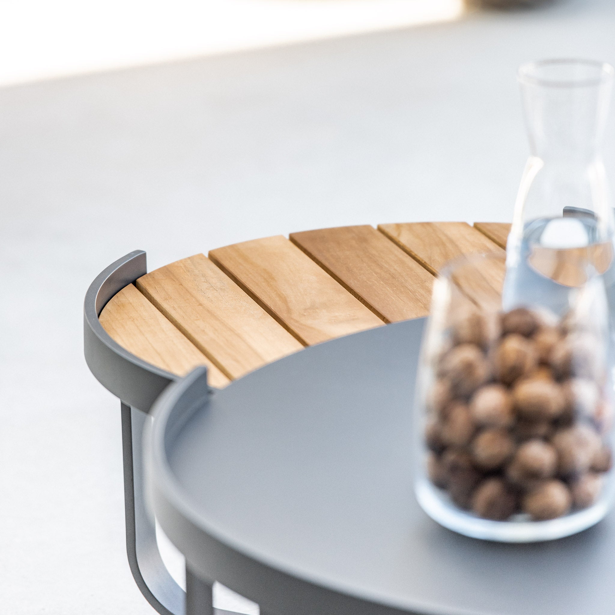 Close-up of the Bloom Coffee Table with Teak Top in Charcoal, showcasing a wooden slat surface and glass jars—one filled with nuts and the other empty—perfect outdoor furniture for your patio.