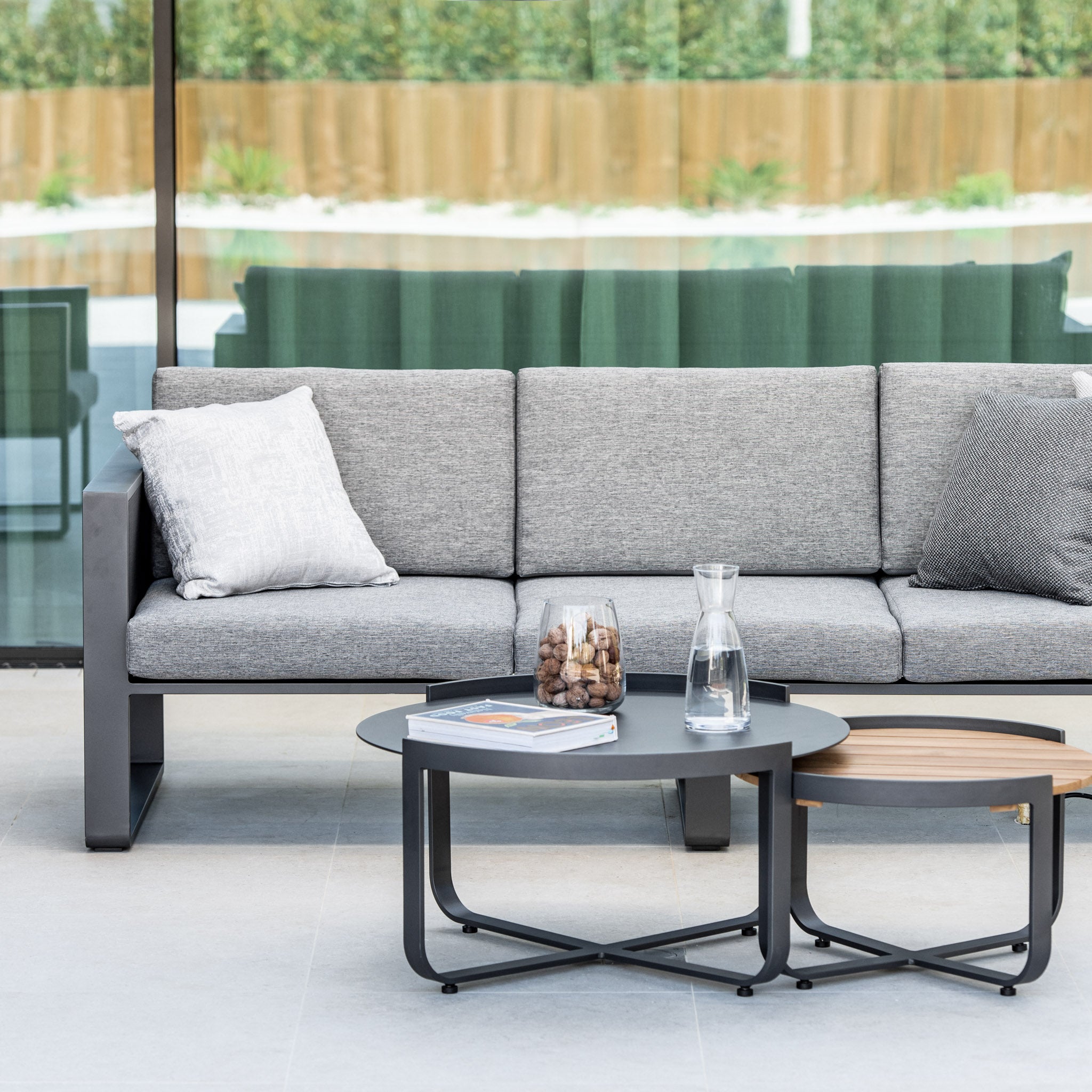 Modern patio with a gray sofa and the Bloom Coffee Table 80cm with Ceramic Top in Charcoal. Small round coffee tables hold a glass pitcher, magazines, and a glass container, completing the look of chic outdoor furniture.