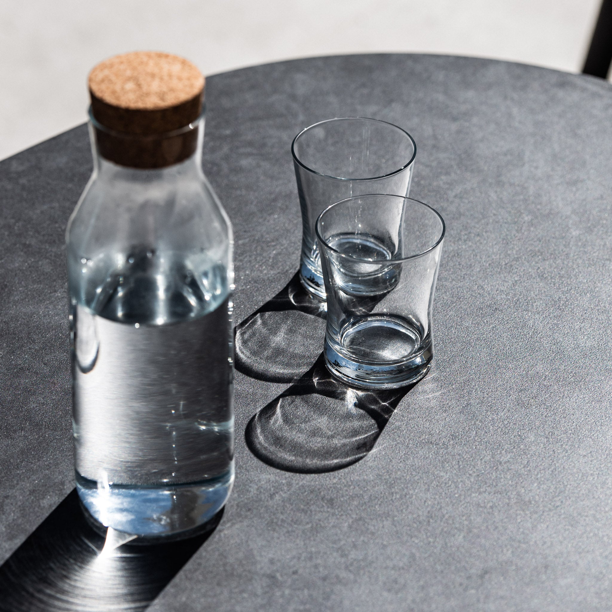 A bottle with a cork lid and two empty glasses casting shadows on a black surface in bright light, perfectly complementing the elegant simplicity of the Cloverly 6 Seat Rope Oval Dining Set with Ceramic Table in Charcoal.