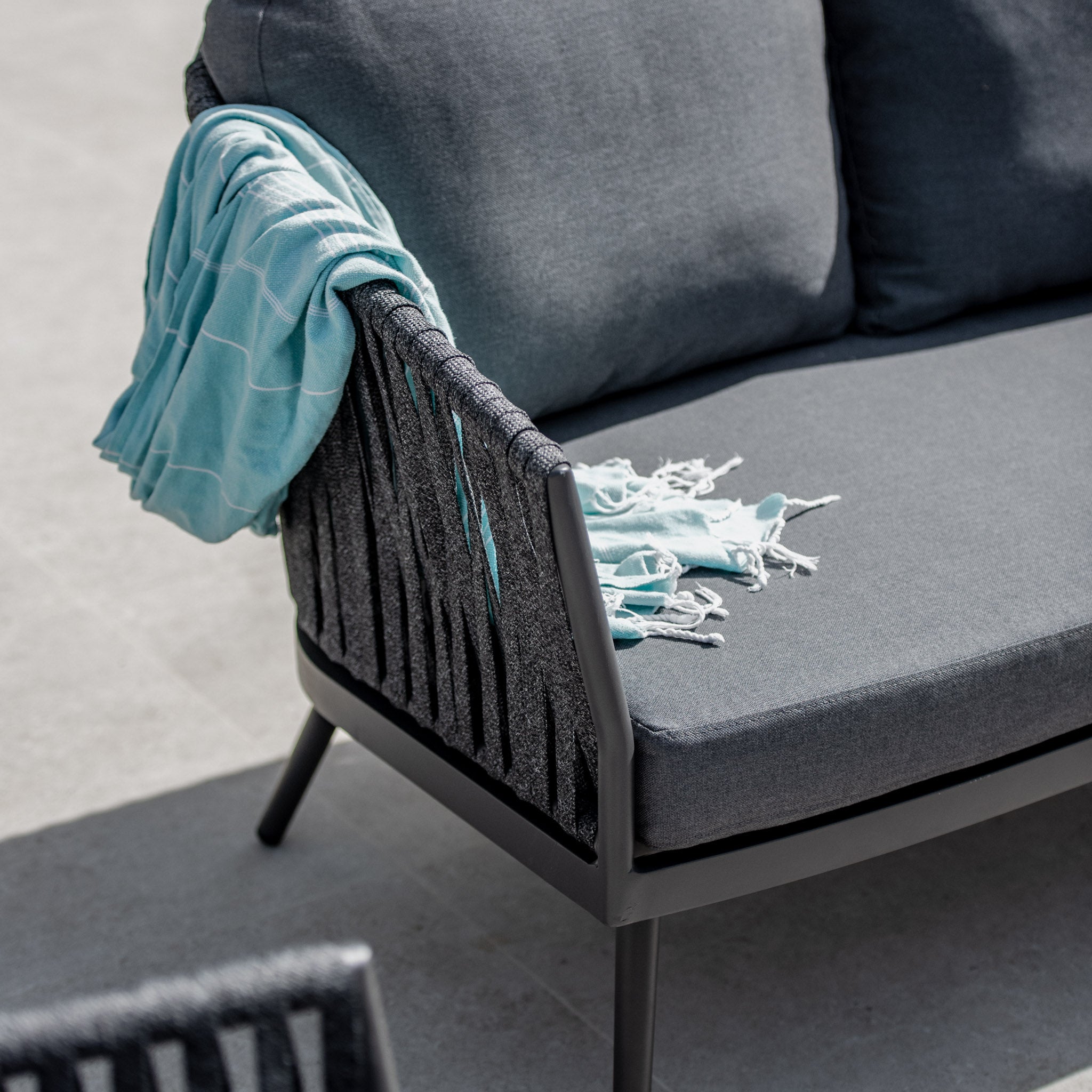 A Monterrey 3 Seat Rope Sofa Set with Rising Firepit Table in Grey, featuring gray cushions and black wicker accents, is adorned with a blue blanket draped over the back, creating a cozy retreat on the light-colored patio.