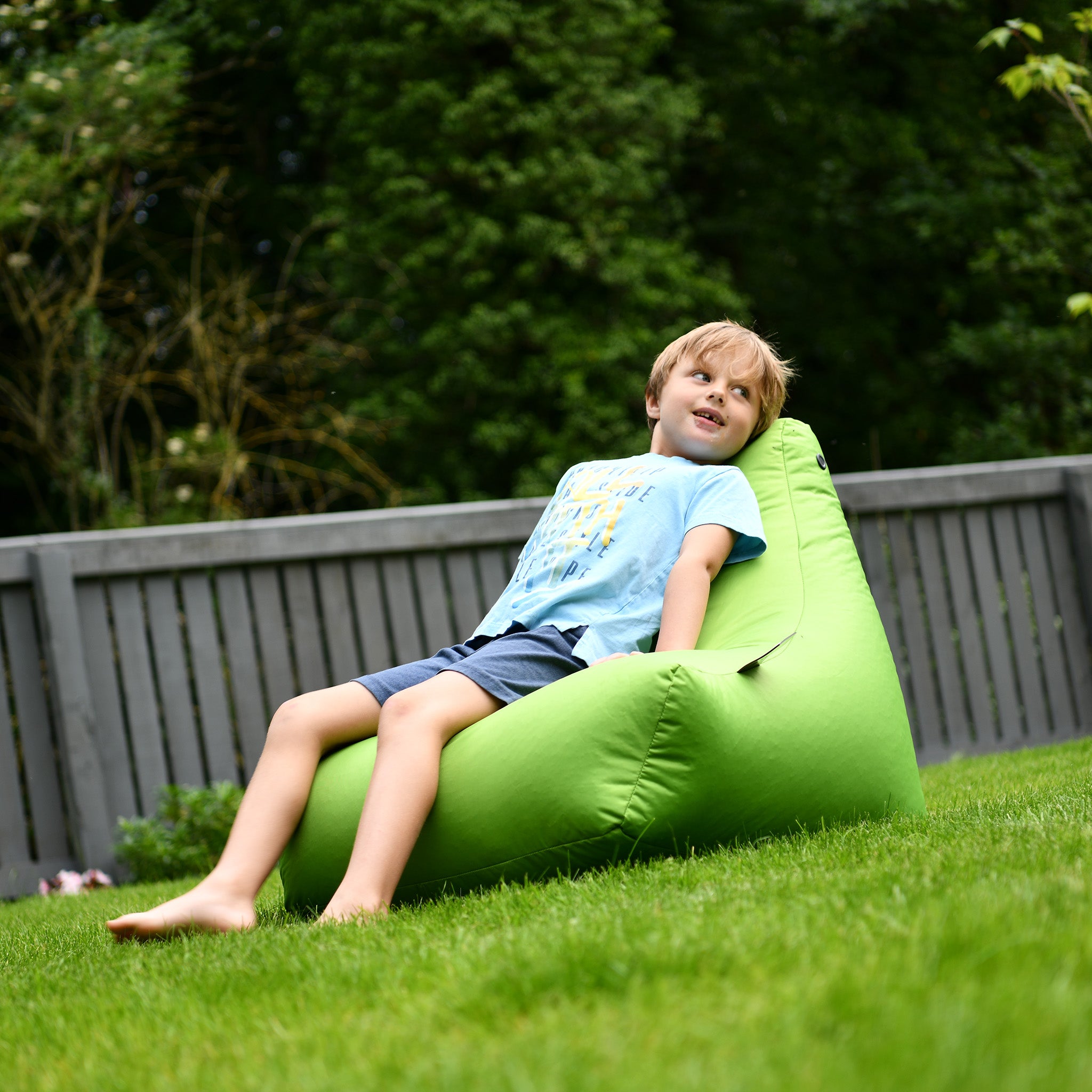 A boy relaxes on an Outdoor Mini B-Bag in Lime, featuring waterproof fabric, in a garden with grass and a gray wooden fence in the background.