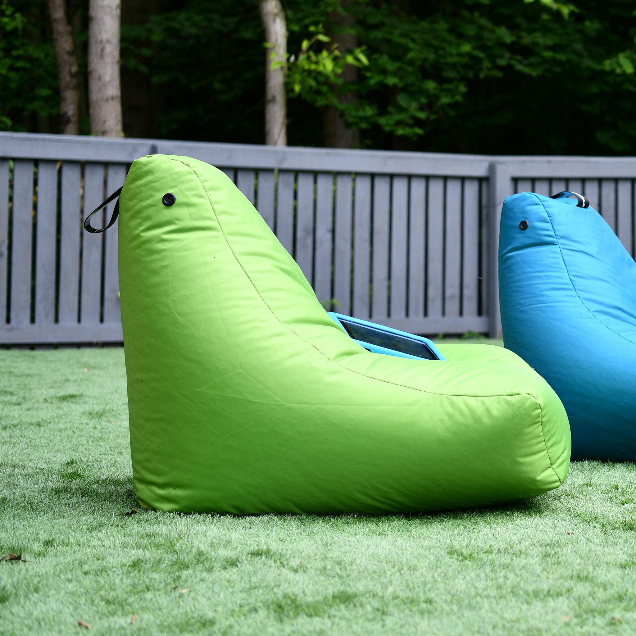 Two Outdoor Mini B-Bags in Lime and Blue sit on grass in front of a grey fence with trees in the background. They're crafted from waterproof fabric, ensuring durability and style come rain or shine.