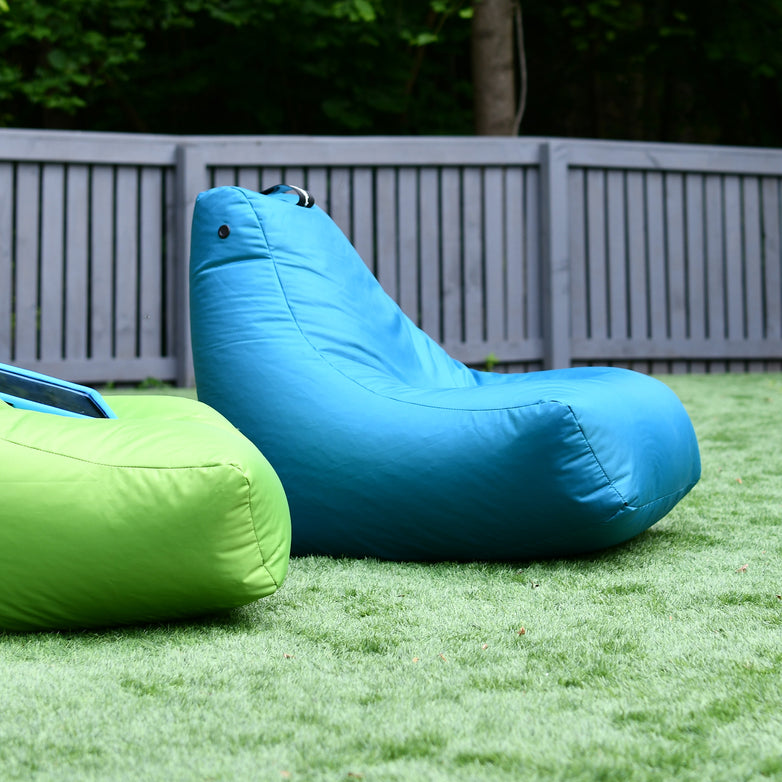 Two colorful Outdoor Mini B-Bags, one in green and one in Aqua, set on a grass lawn with a grey wooden fence in the background. Crafted from waterproof fabric and fade-resistant, these chairs are perfect for lounging in any weather.