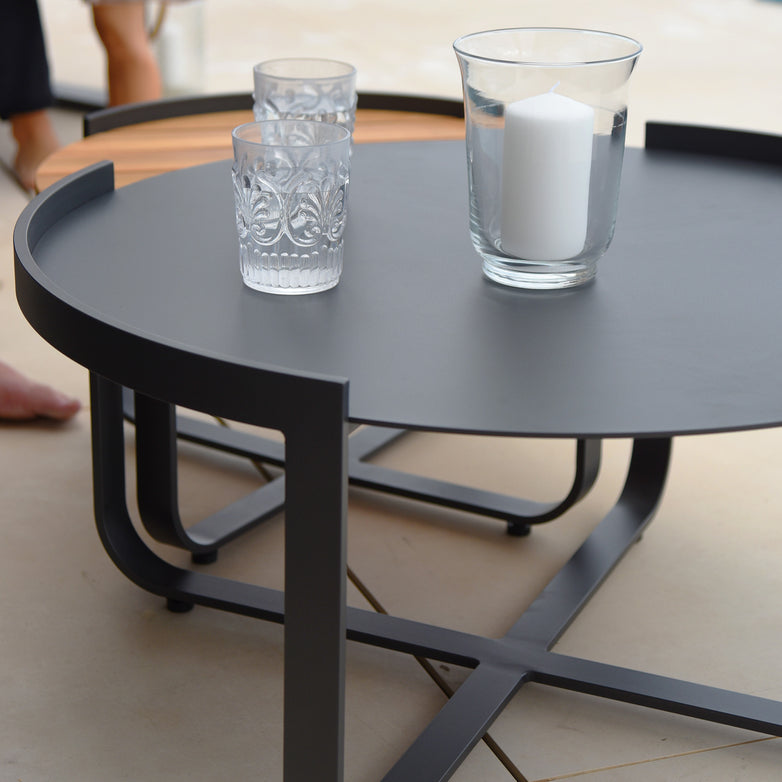 The Bloom Coffee Table 80cm with Ceramic Top in Charcoal, with its round black design, features a ceramic glass top adorned with a glass candle holder and two patterned drinking glasses.