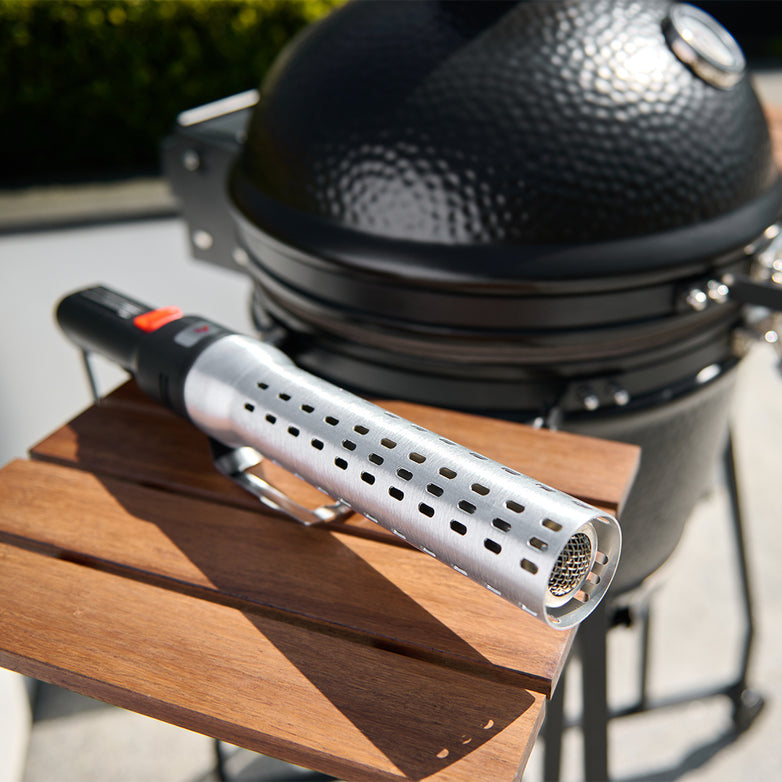An Electric Charcoal Lighter for Kamados & BBQ Grills with a perforated design rests on a wooden table beside a barbecue grill, ideal for igniting superheated air in charcoal BBQs.