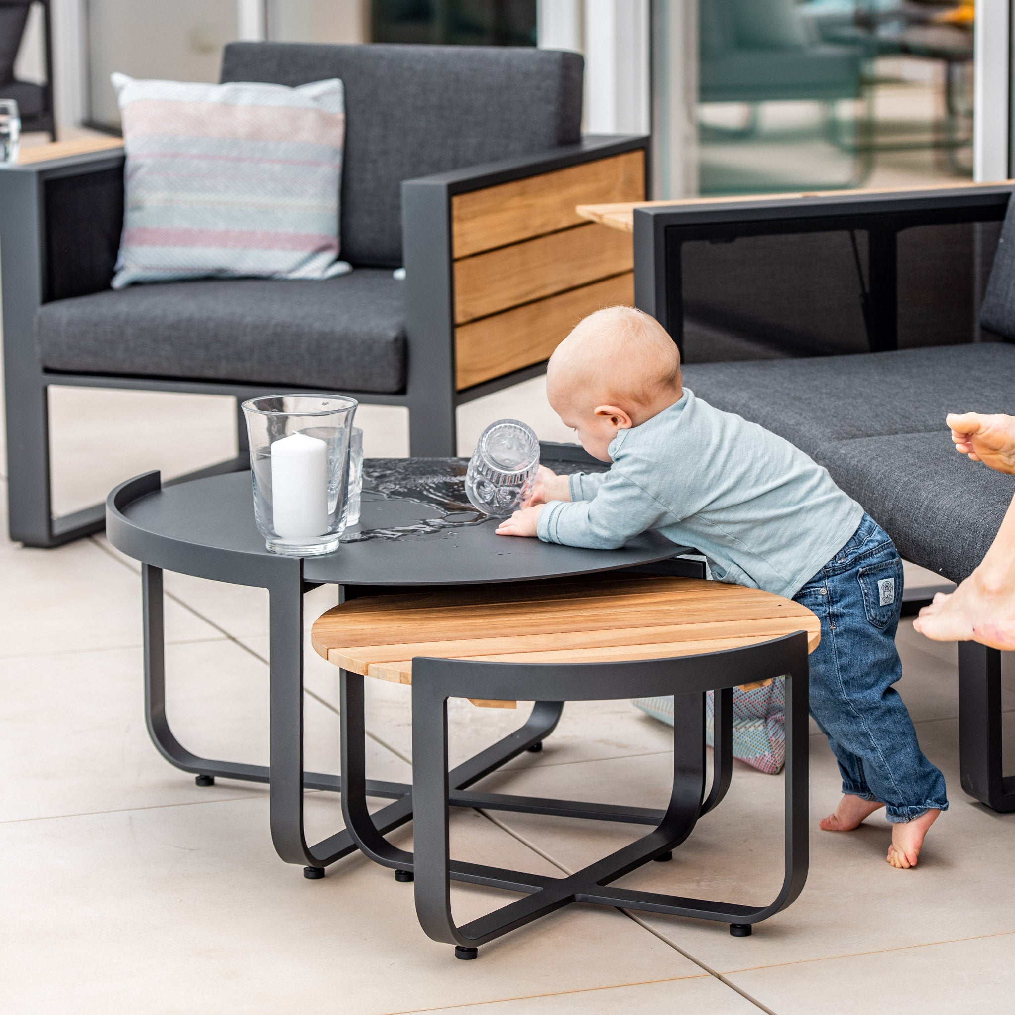 The baby stands and plays on the Bloom Coffee Table 80cm with Ceramic Top in Charcoal, next to a spilled glass cup. Modern outdoor furniture featuring ceramic glass tops in the background enhances the stylish scene.