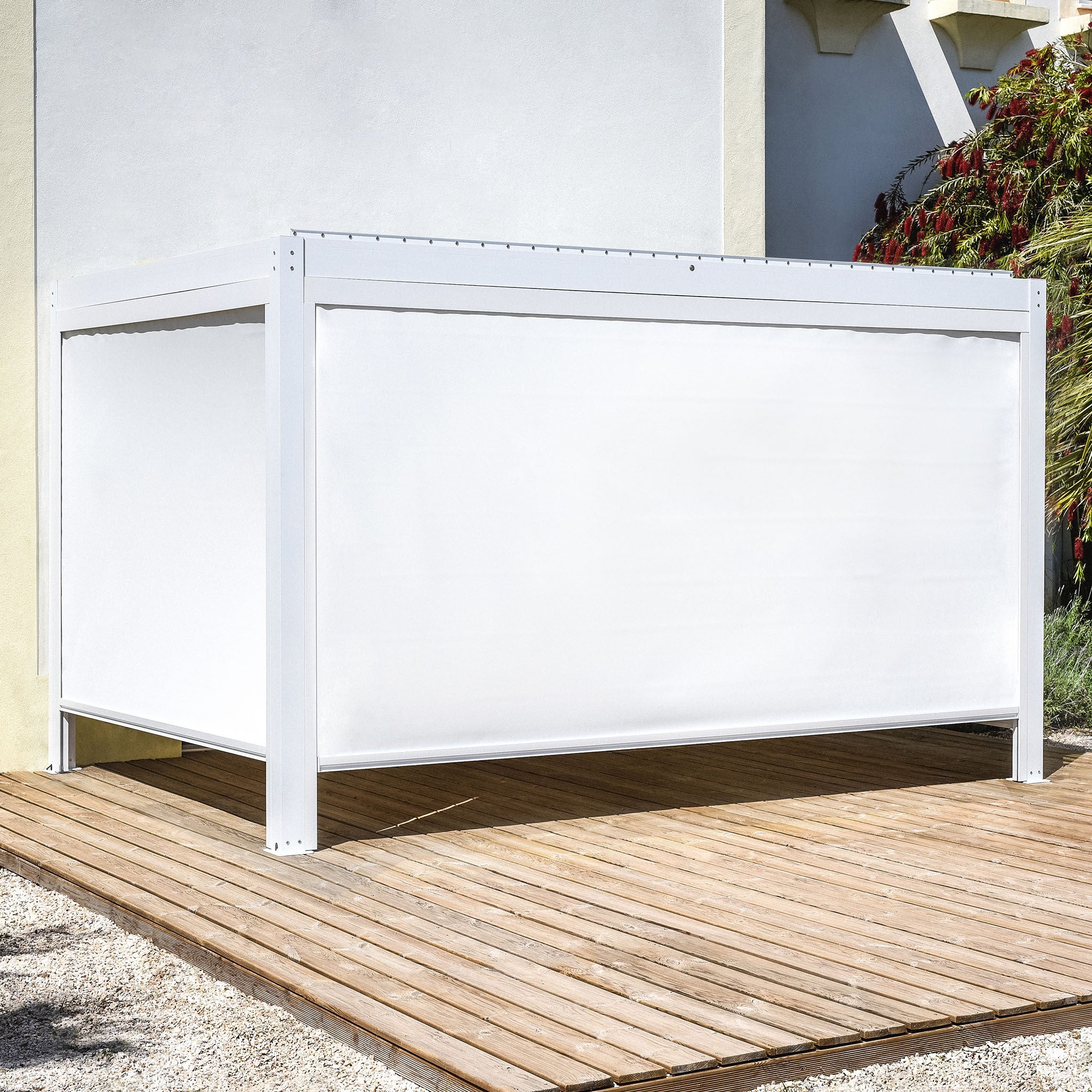 A sleek white cube structure on a wooden deck, featuring the PergoSTET Plus 3m x 4m Rectangular Pergola with 3 drop sides and LED lighting in white, stands gracefully beside a building adorned with lush green foliage.