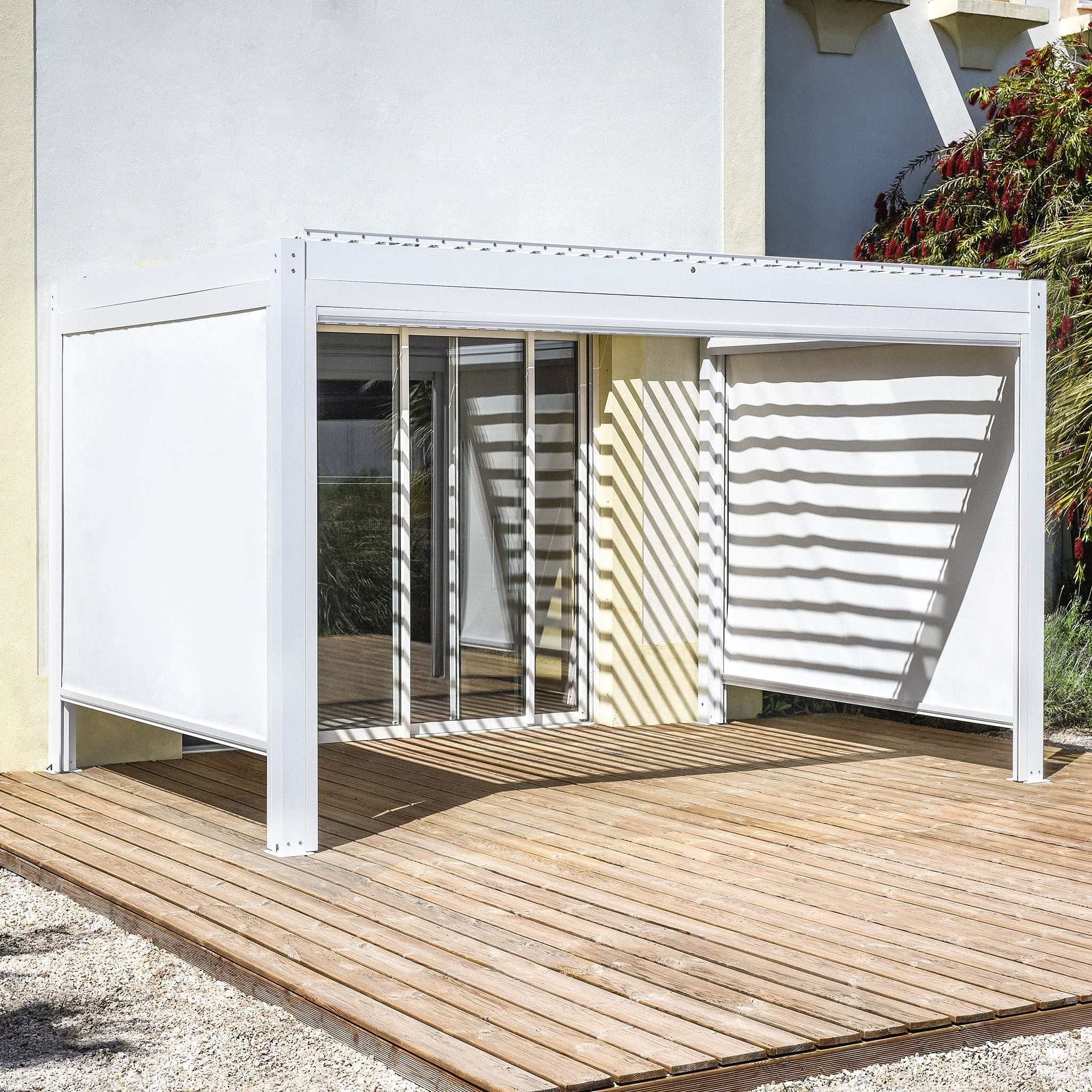 The sleek PergoSTET Plus 3m x 4m rectangular pergola with white horizontal slats and LED lighting casts intricate shadows on the wooden deck by the glass door, featuring a waterproof roof that ensures all-weather enjoyment and includes three drop sides for added protection.