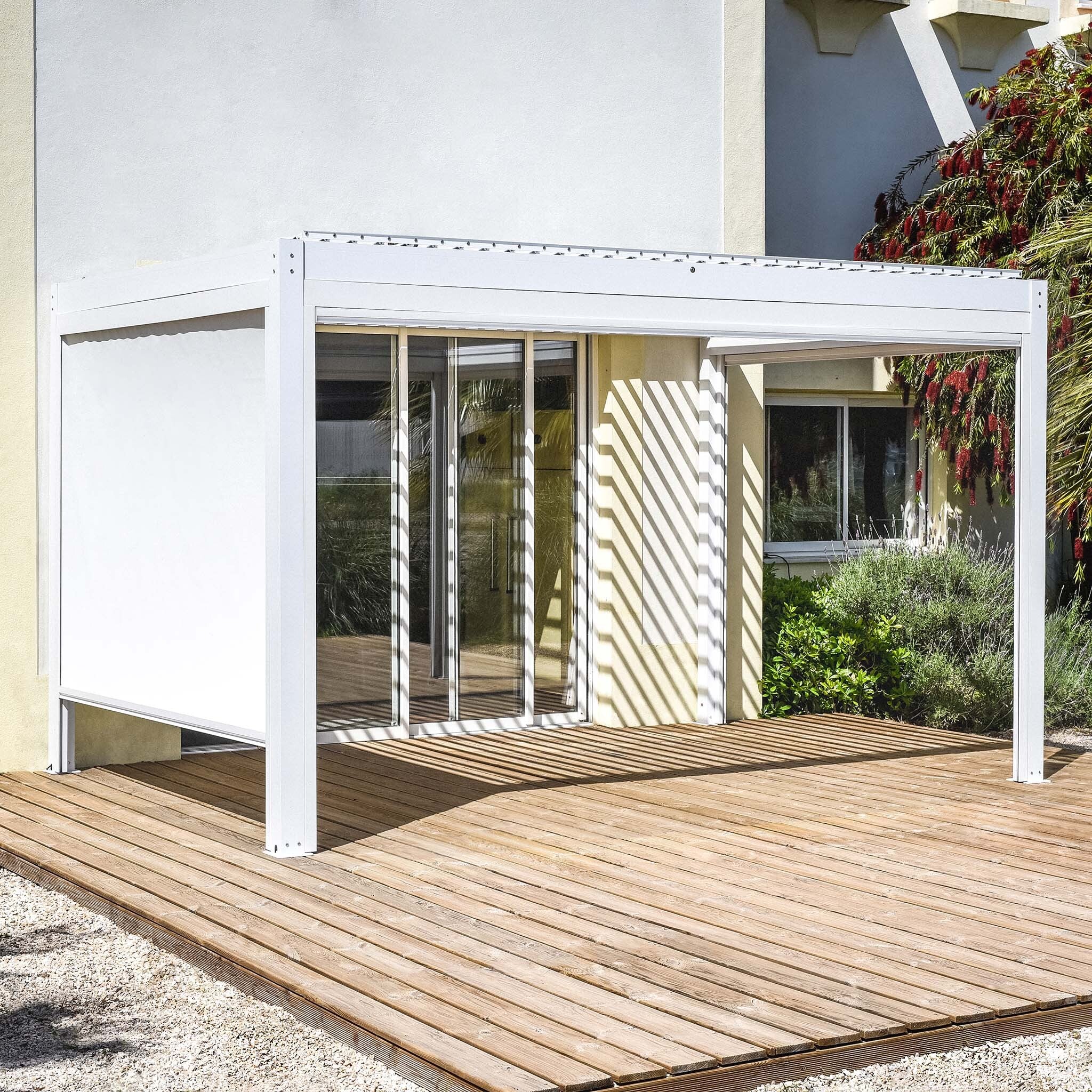 The PergoSTET Plus 3m x 4m Rectangular Pergola with 3 Drop Sides and LED Lighting in White enhances a wooden deck outside a contemporary home. Its waterproof roof and built-in drainage system provide comfort in all weather, while sliding glass doors and lush plants complete the stylish design.