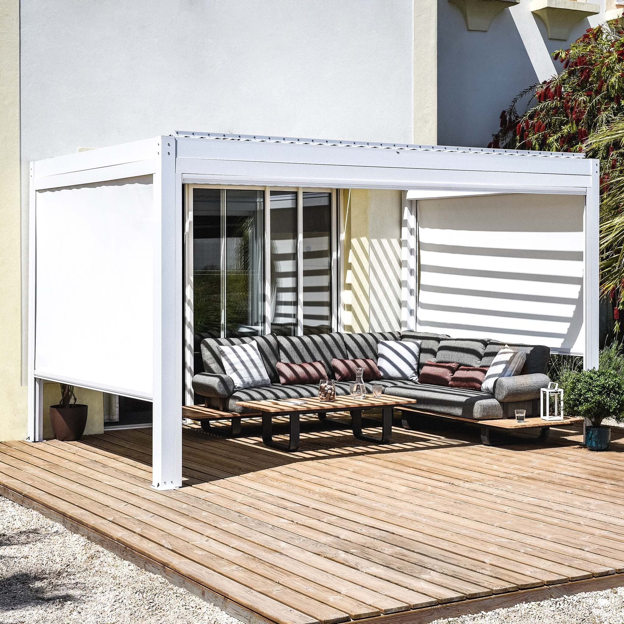 A modern patio showcases a white PergoSTET Plus 3m x 4m rectangular pergola with LED lighting and drop sides, an L-shaped sofa with striped cushions, a wooden deck, and lush plants. Shadow patterns dance across the space, adding to its charm.