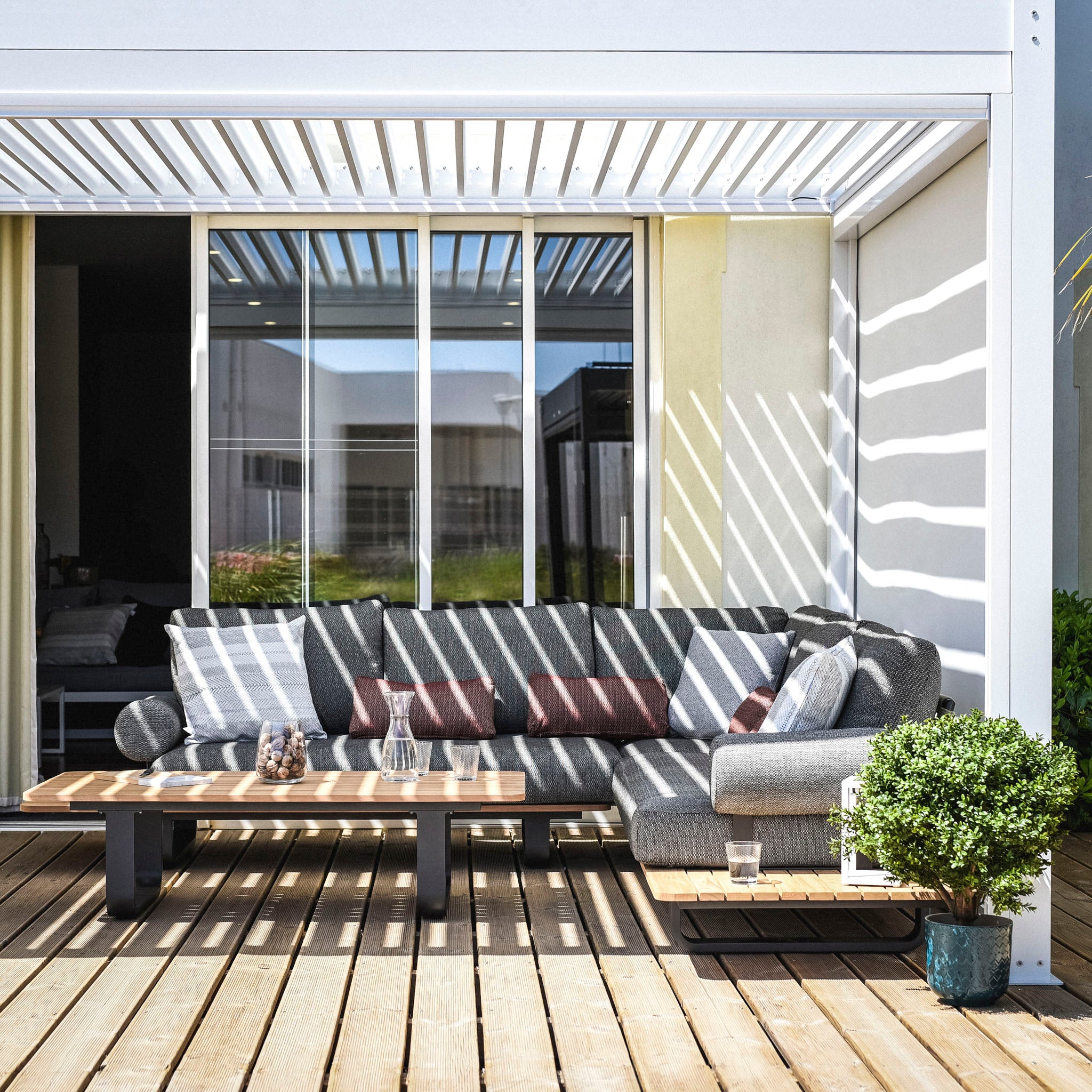 Outdoor patio with a gray sofa, shadows from a PergoSTET Plus 3m x 4m Rectangular Pergola with LED lighting, wooden deck, and small table with a plant.