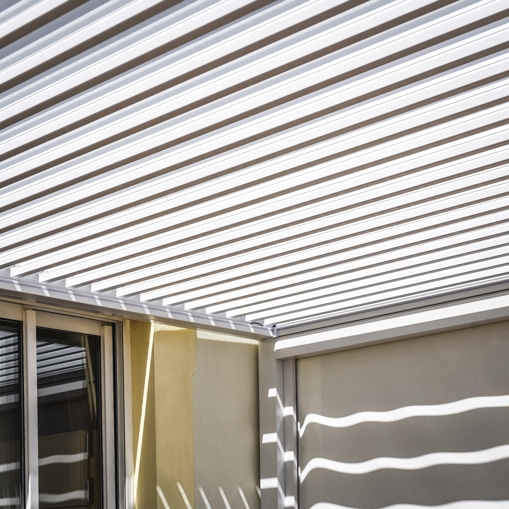 The PergoSTET Plus 3m x 4m Rectangular Pergola in White features a slatted roof casting striped shadows and a built-in drainage system, embodying sleek modern design with 3 drop sides and LED lighting.