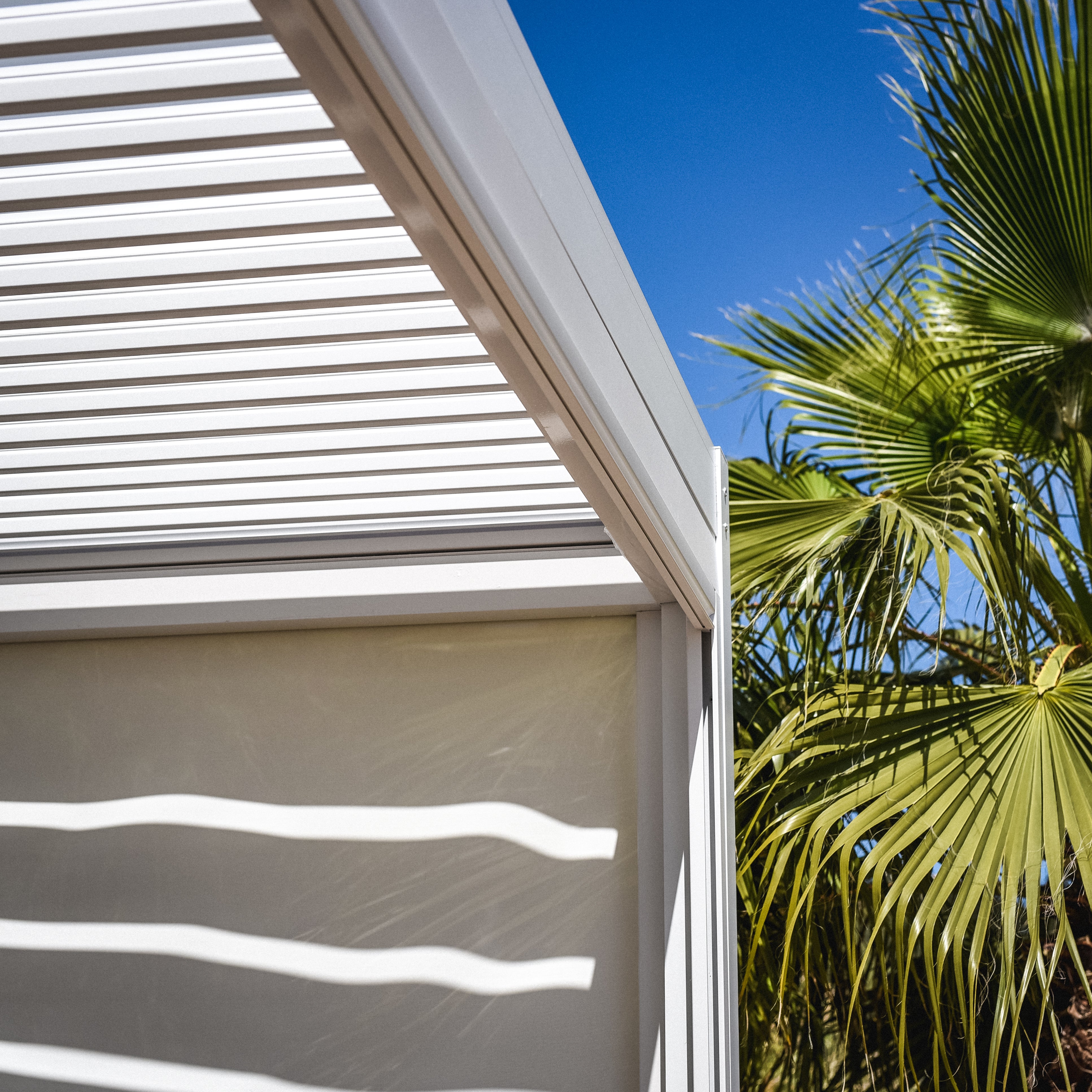 The PergoSTET Plus 4m x 4m Square Pergola with LED Lighting (white frame only) casts striped shadows, with a green palm tree and clear blue sky in the background.