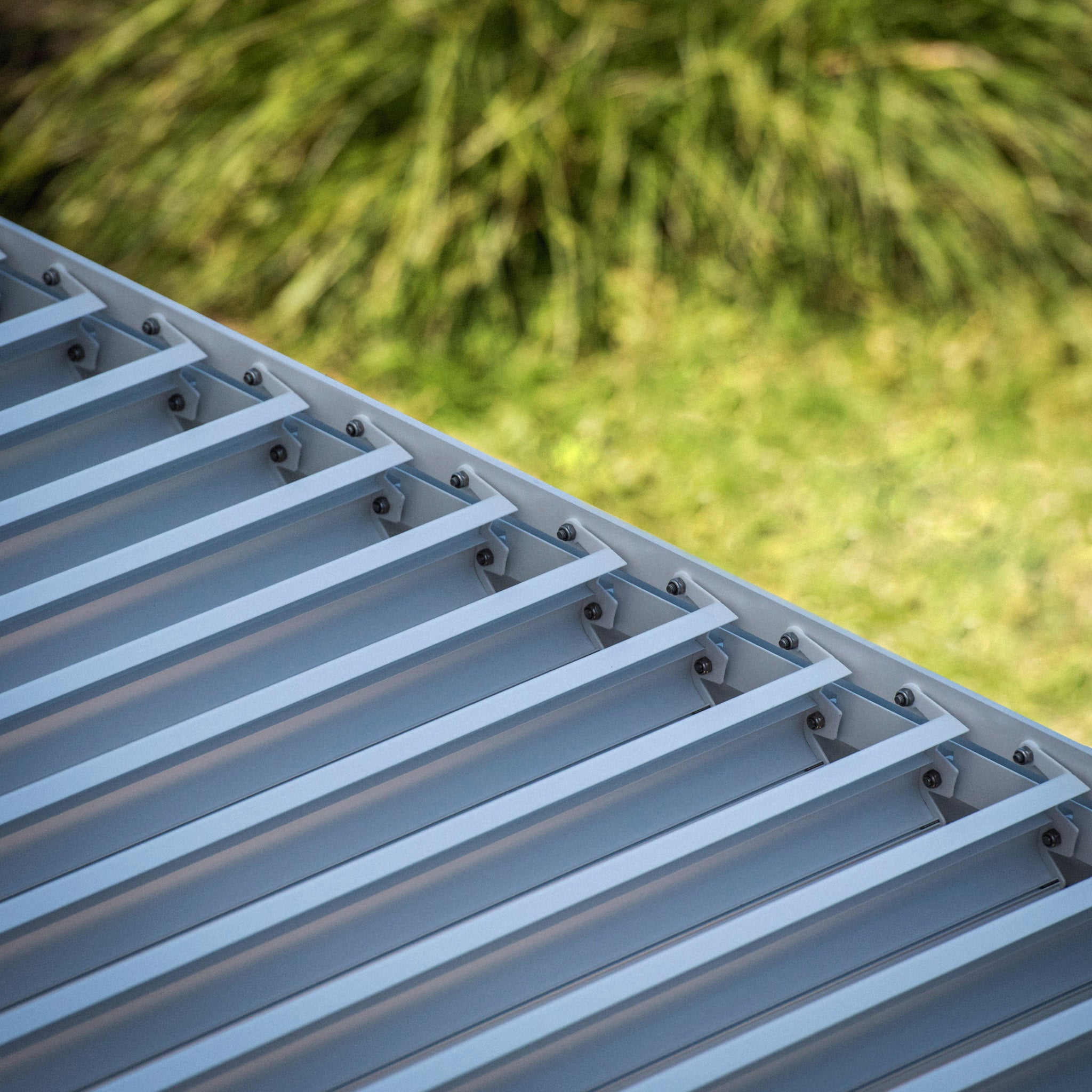 The PergoSTET Plus Pergola with LED lighting features visible screws on slanted metal roof panels and a built-in drainage system, set against a blurred green grass background.