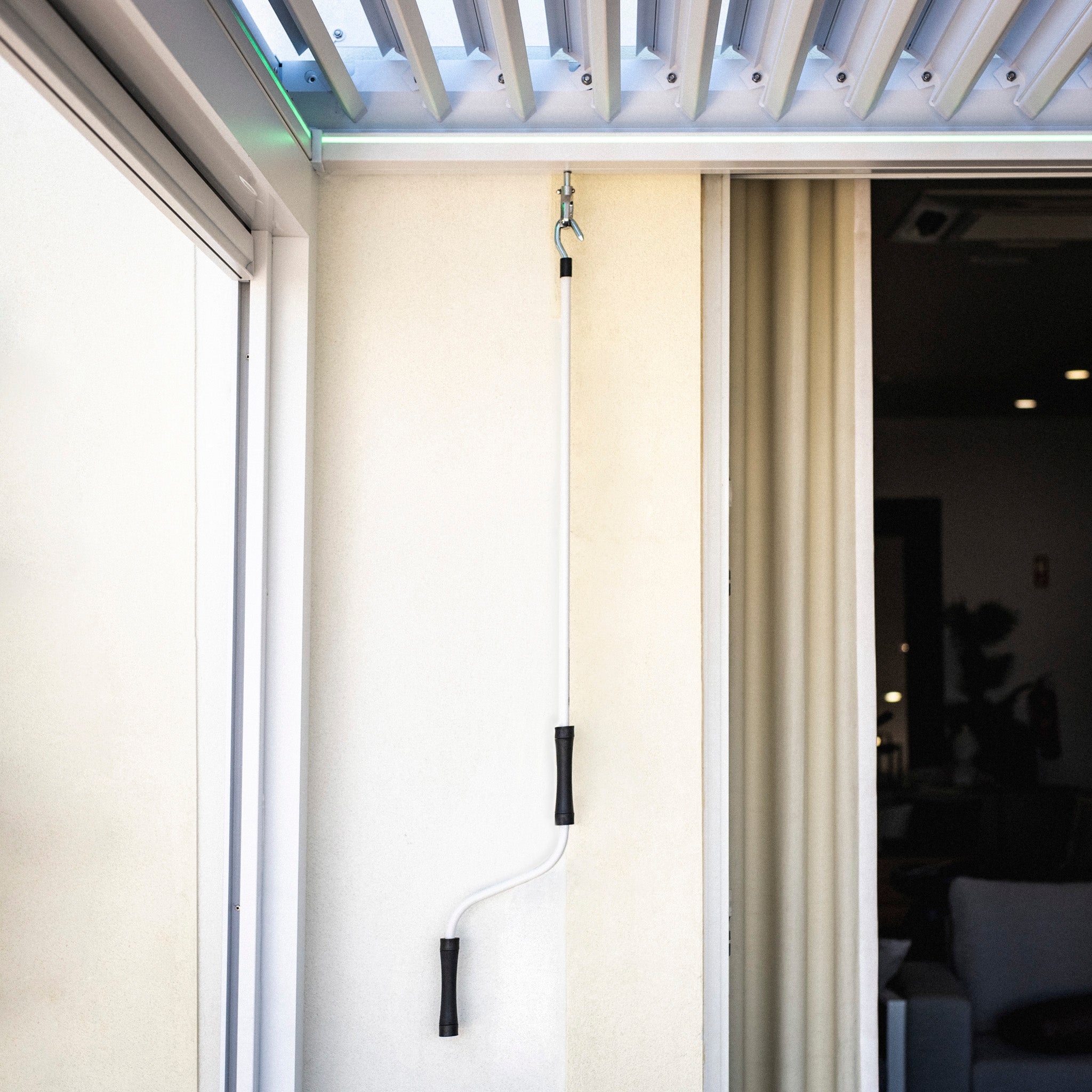 A long white adjustment pole with black handles, hanging beside a sliding door and curtains, complements the sleek design of the PergoSTET Plus 3m x 4m Rectangular Pergola with Drop Sides and LED Lighting in White, featuring a built-in drainage system.