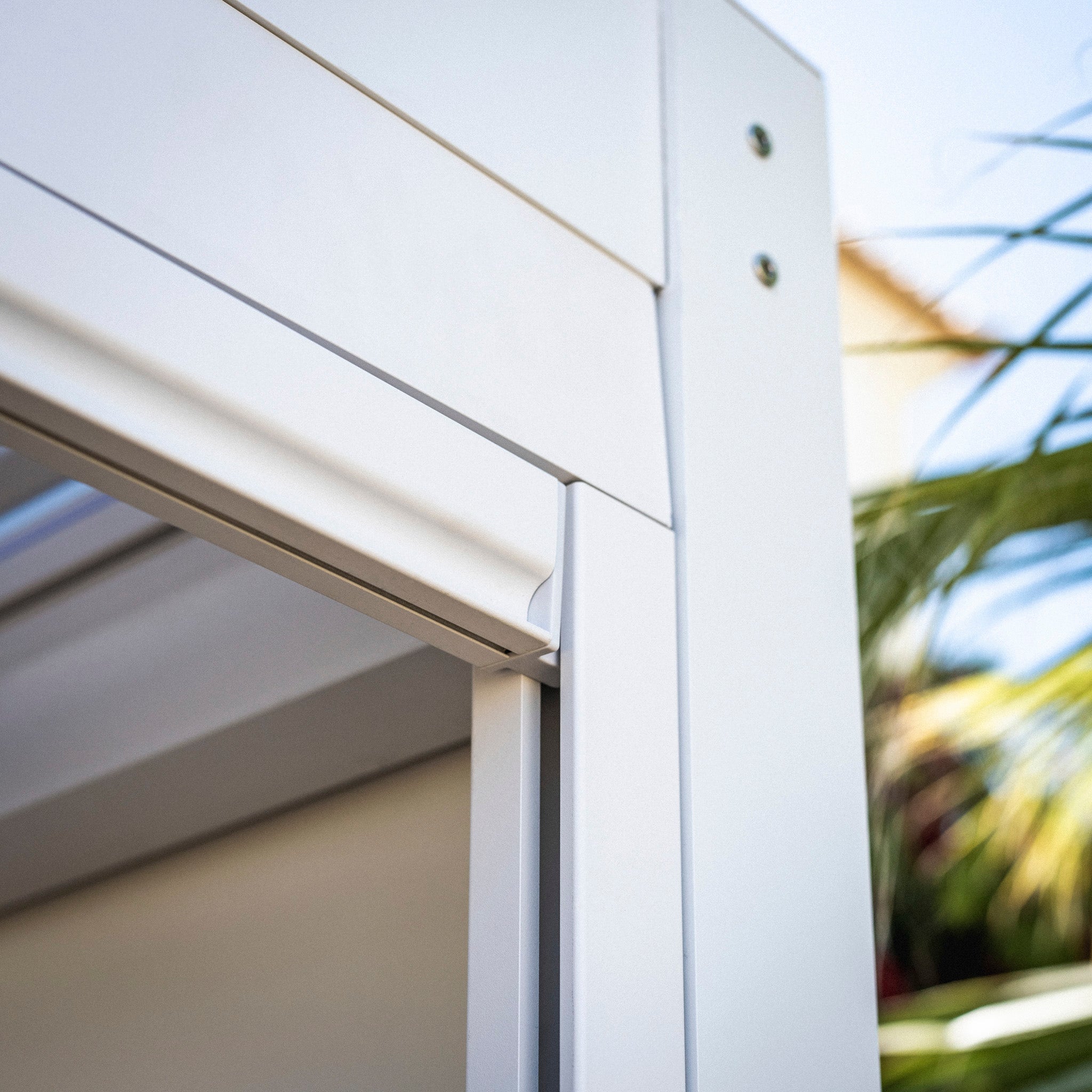Close-up of a PergoSTET Plus 3m x 4m rectangular pergola in white, featuring palm leaves blurred in the background, showcased with innovative built-in drainage and LED lighting.