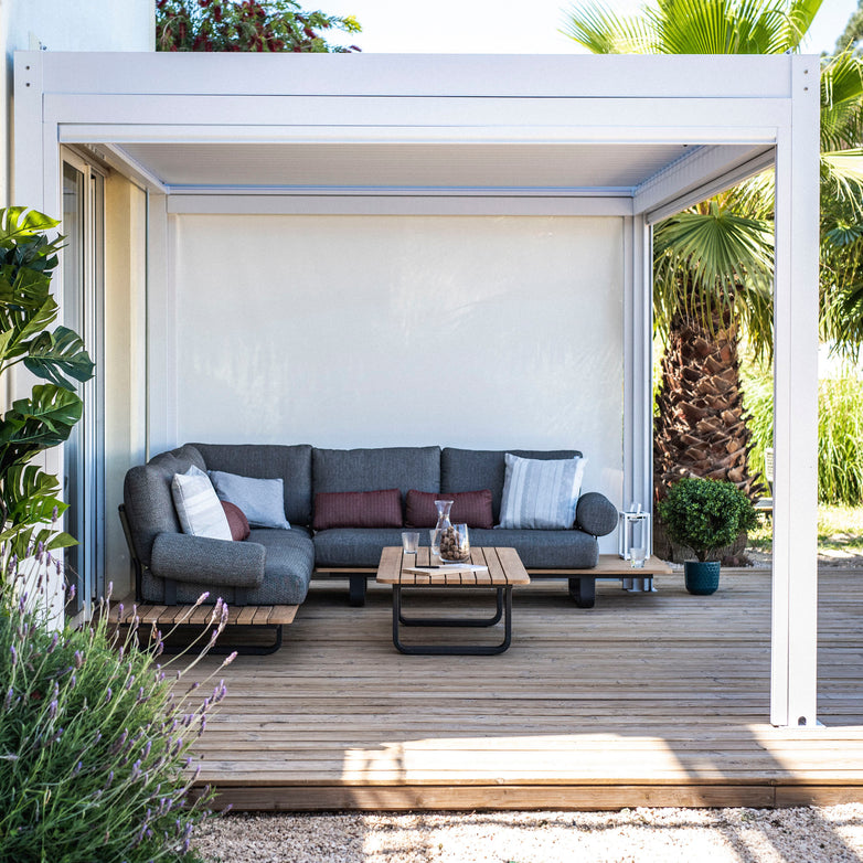 A modern patio showcases a gray sectional, wooden coffee table, and plants beneath a PergoSTET Plus Pergola. This innovative 3m x 3m design includes waterproof roofing, LED lighting, and drop sides for all-weather comfort and style.