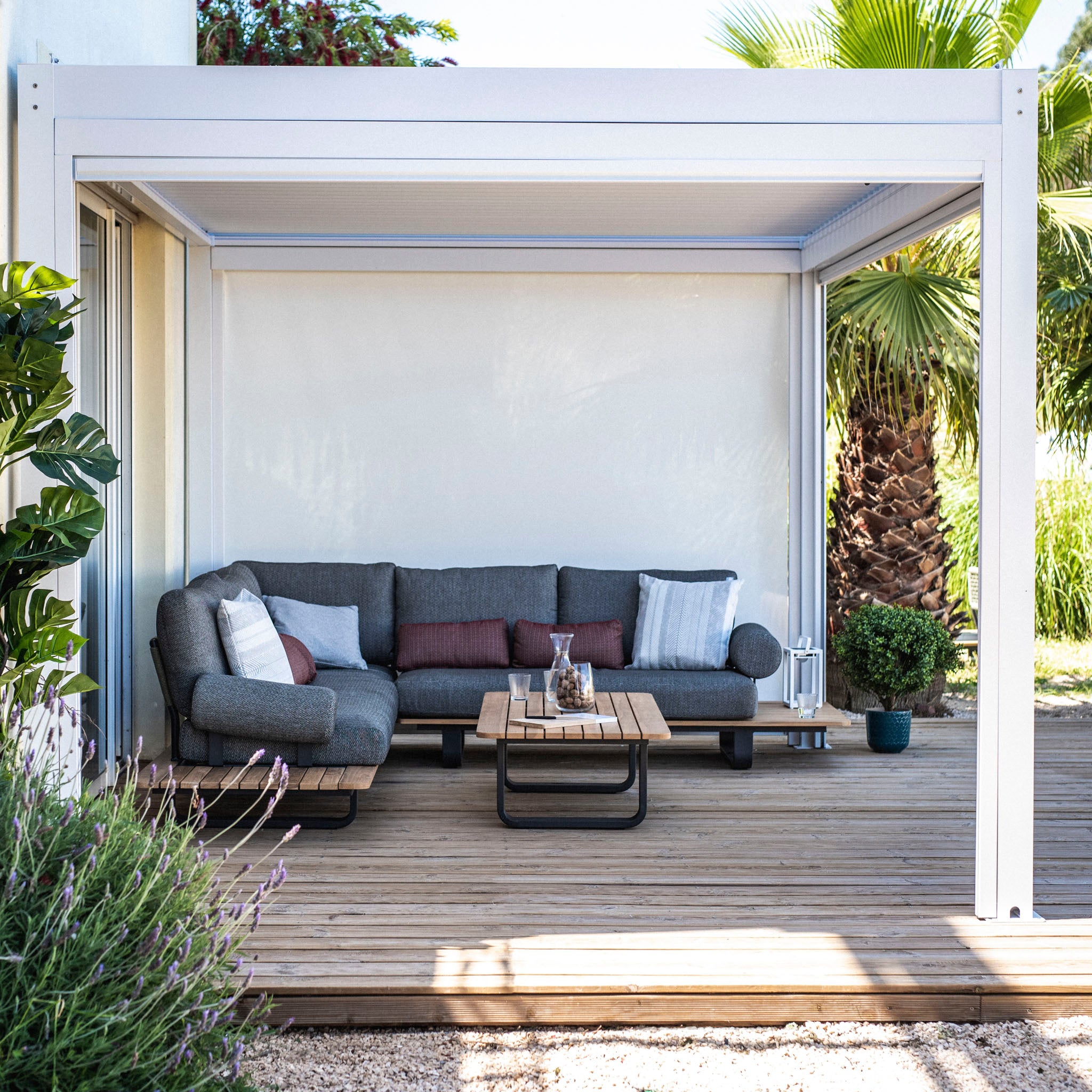 A modern patio features a gray sectional sofa, wooden coffee table, and potted plants under the PergoSTET Plus 3m x 4m Pergola with built-in drainage and LED lighting. The sunny setting perfectly complements this blend of comfort and innovation.