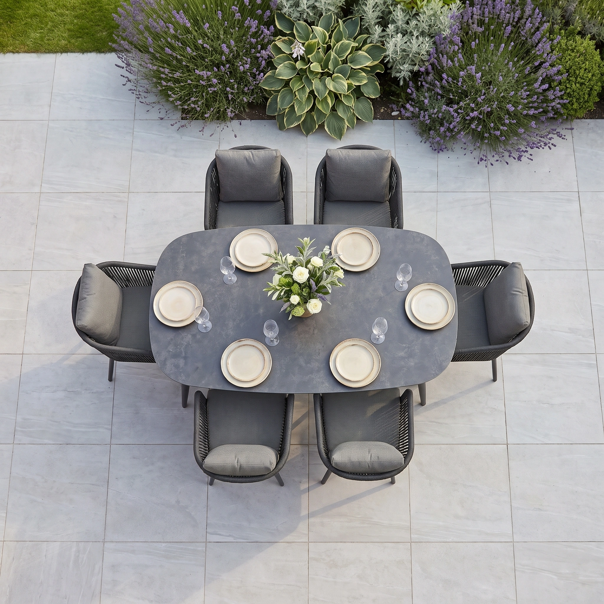The Dekota 6 Seat Dining Set with Albatross Oval Dining Table in Charcoal features a durable sintered stone tabletop and elegant rope dining chairs, complete with place settings and a floral centerpiece, all surrounded by lush garden plants.