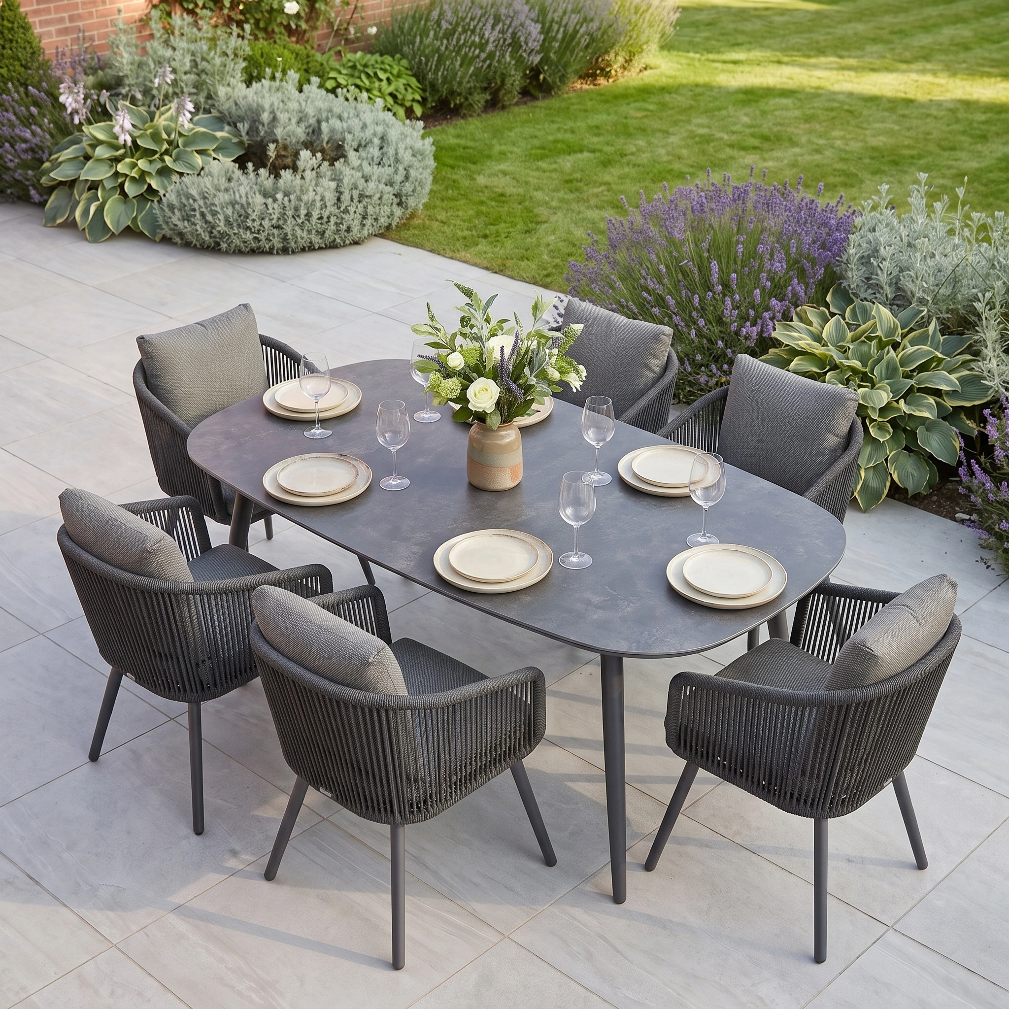 The Dekota 6 Seat Dining Set with Albatross Oval Dining Table in Charcoal features gray rope dining chairs and a durable sintered stone tabletop, complemented by plates, glasses, a floral centerpiece, and surrounded by lush plants.