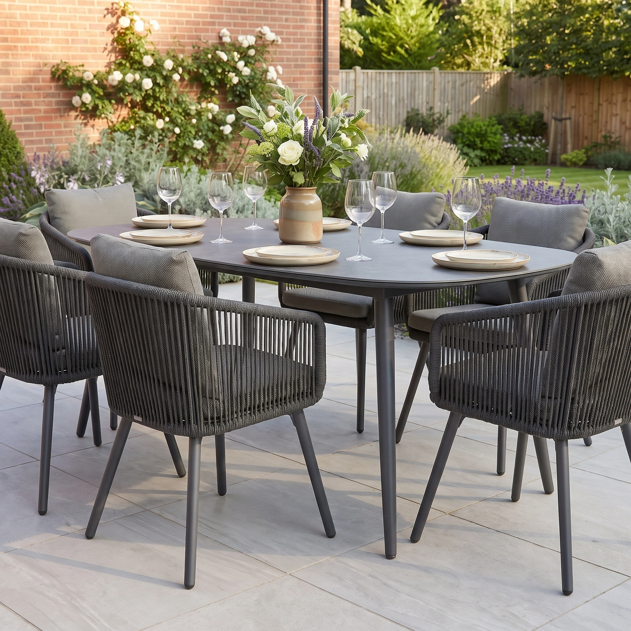 The Dekota 6 Seat Dining Set with Albatross Oval Dining Table in Charcoal features stylish rope dining chairs and a durable sintered stone tabletop, beautifully arranged with plates, glasses, and a flower vase on an inviting patio.