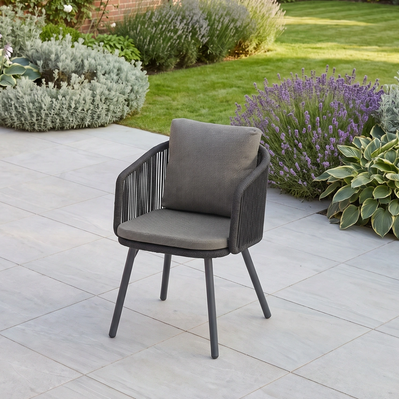 A gray cushioned outdoor chair sits on a patio, surrounded by green plants and purple flowers, perfectly complementing the Dekota 6 Seat Dining Set with Ravona Extending Table in Charcoal for stylish outdoor gatherings.