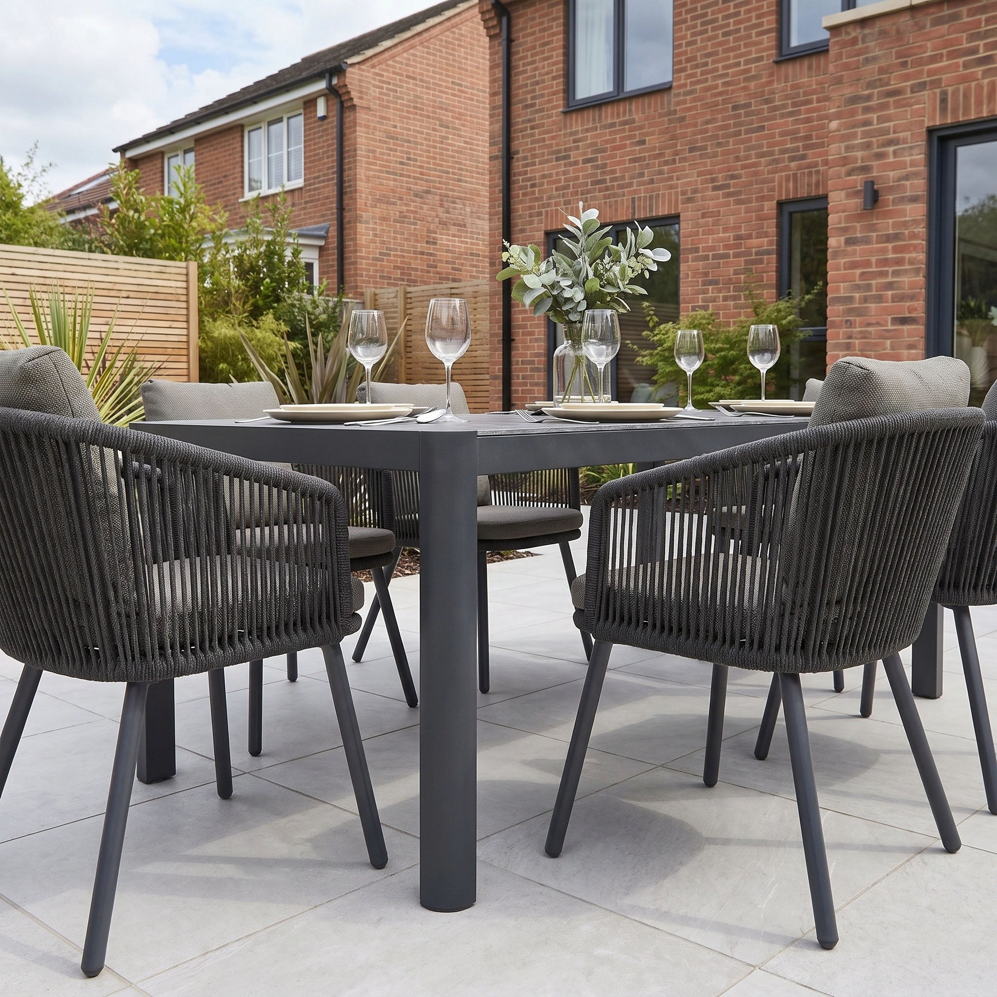 Experience modern outdoor dining with the Dekota 6 Seat Dining Set, featuring stylish Dekota Dining Chairs beautifully arranged around the Ravona Extending Table in Charcoal. Elegantly set with plates, glasses, and a vase, this contemporary set combines comfort and sophistication—perfect for elevating your al fresco gatherings.