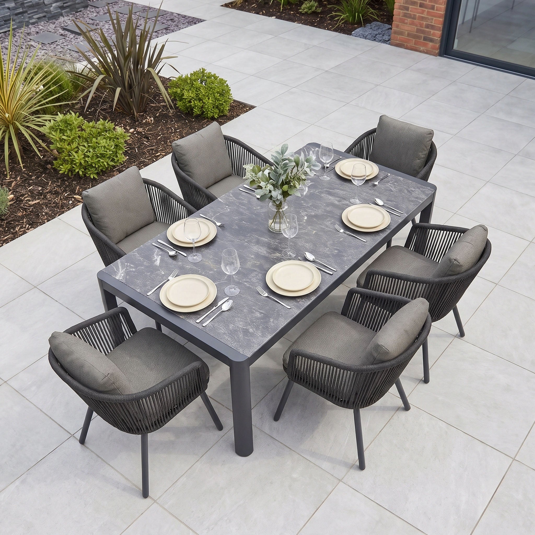 The Dekota 6 Seat Dining Set with Ravona Extending Table in Charcoal features an elegant aluminium extending table paired with six plush-cushioned Dekota Dining Chairs, beautifully arranged with plates and glasses on a tiled patio.