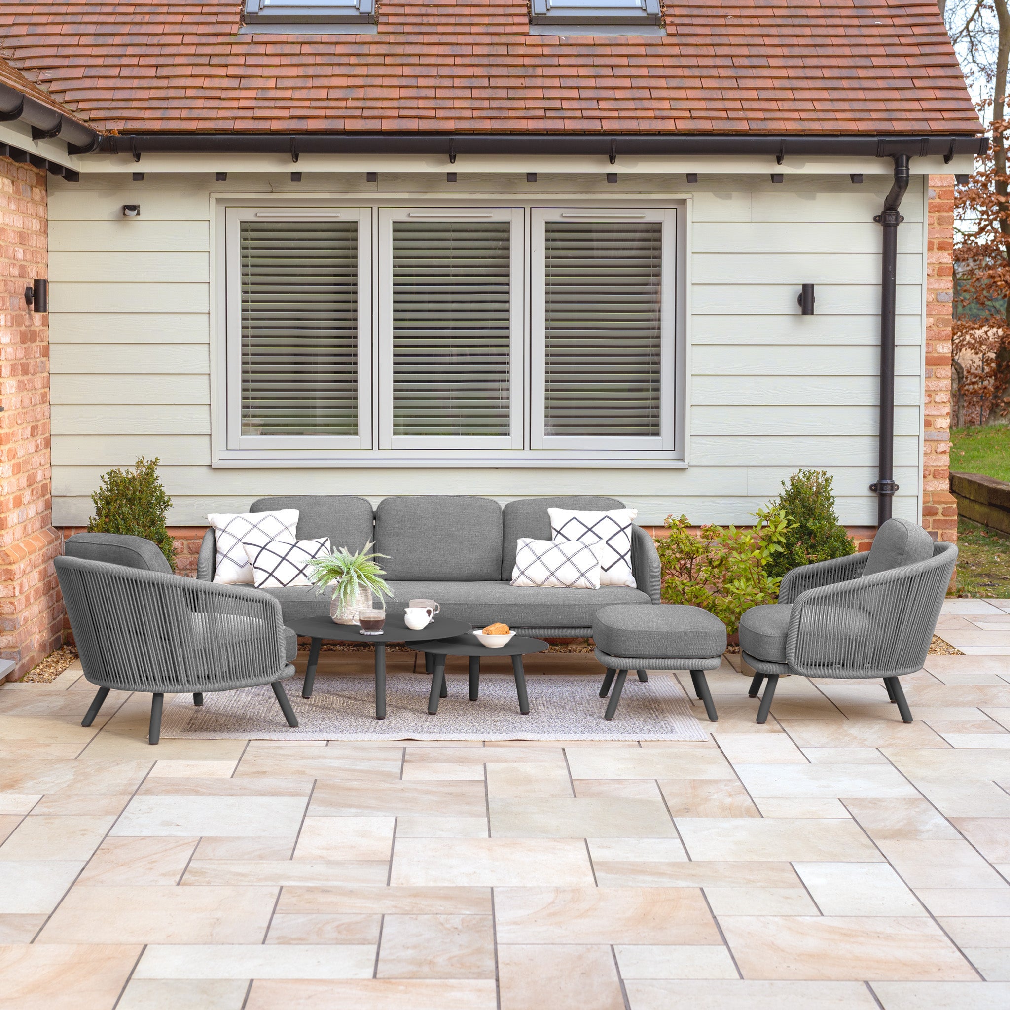 Charcoal Dekota 3 Seat Rope Sofa Set with coffee tables and a footstool, crafted with durable materials, is arranged on a stone-tiled patio in front of a house.